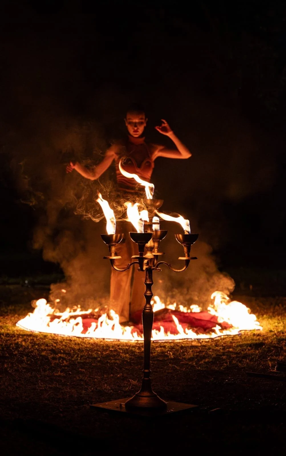 Une personne dans un spectacle de feu, se tenant derrière un chandelier allumé, avec un feu au sol et dansant avec des flammes.