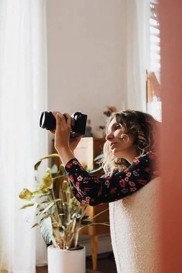 Femme assise près d'une fenêtre, tenant un appareil photo, regardant par la fenêtre.