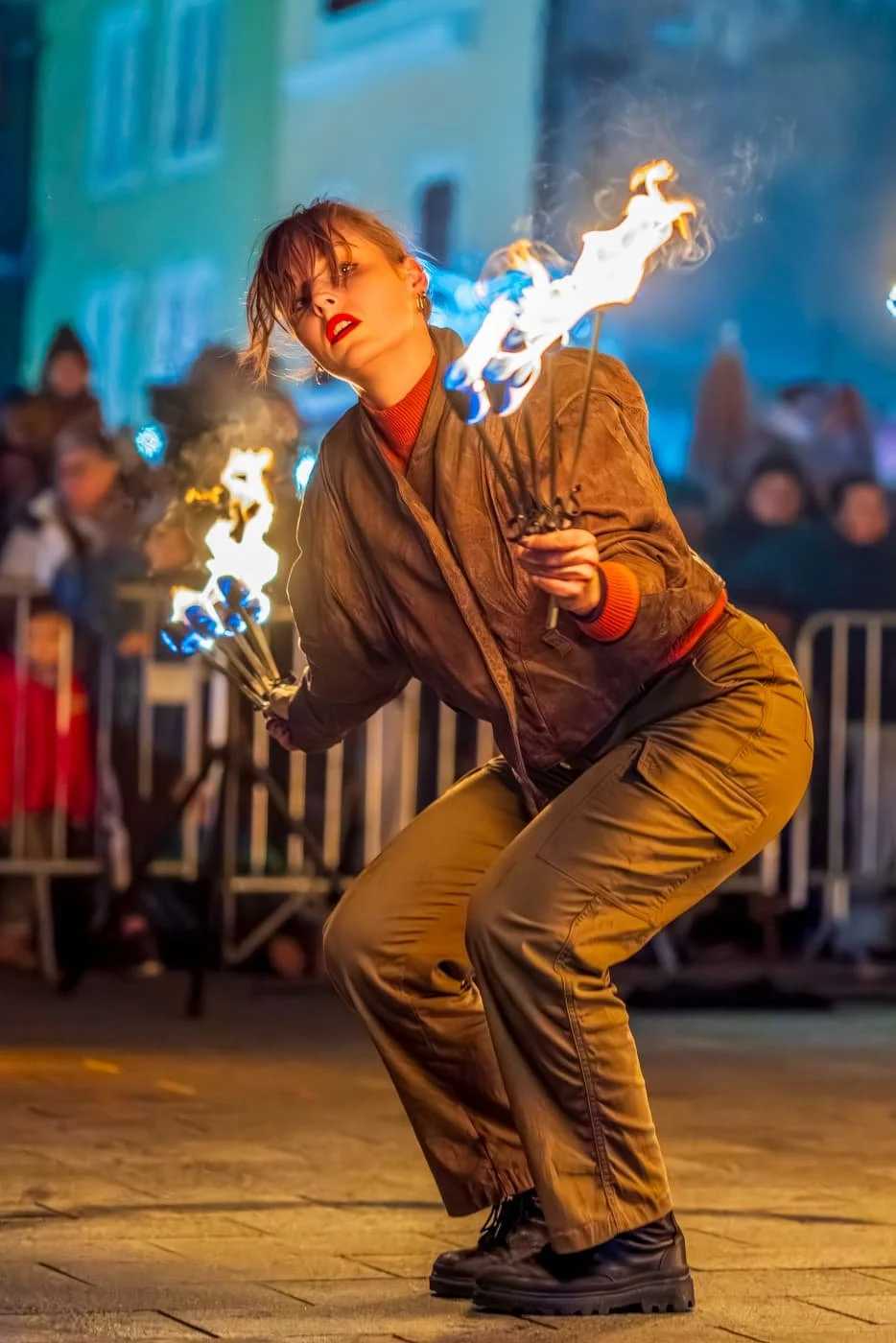 Une femme en costume danse dans une rue, tenant deux torches enflammées, lors d'une performance nocturne avec un public en arrière-plan.