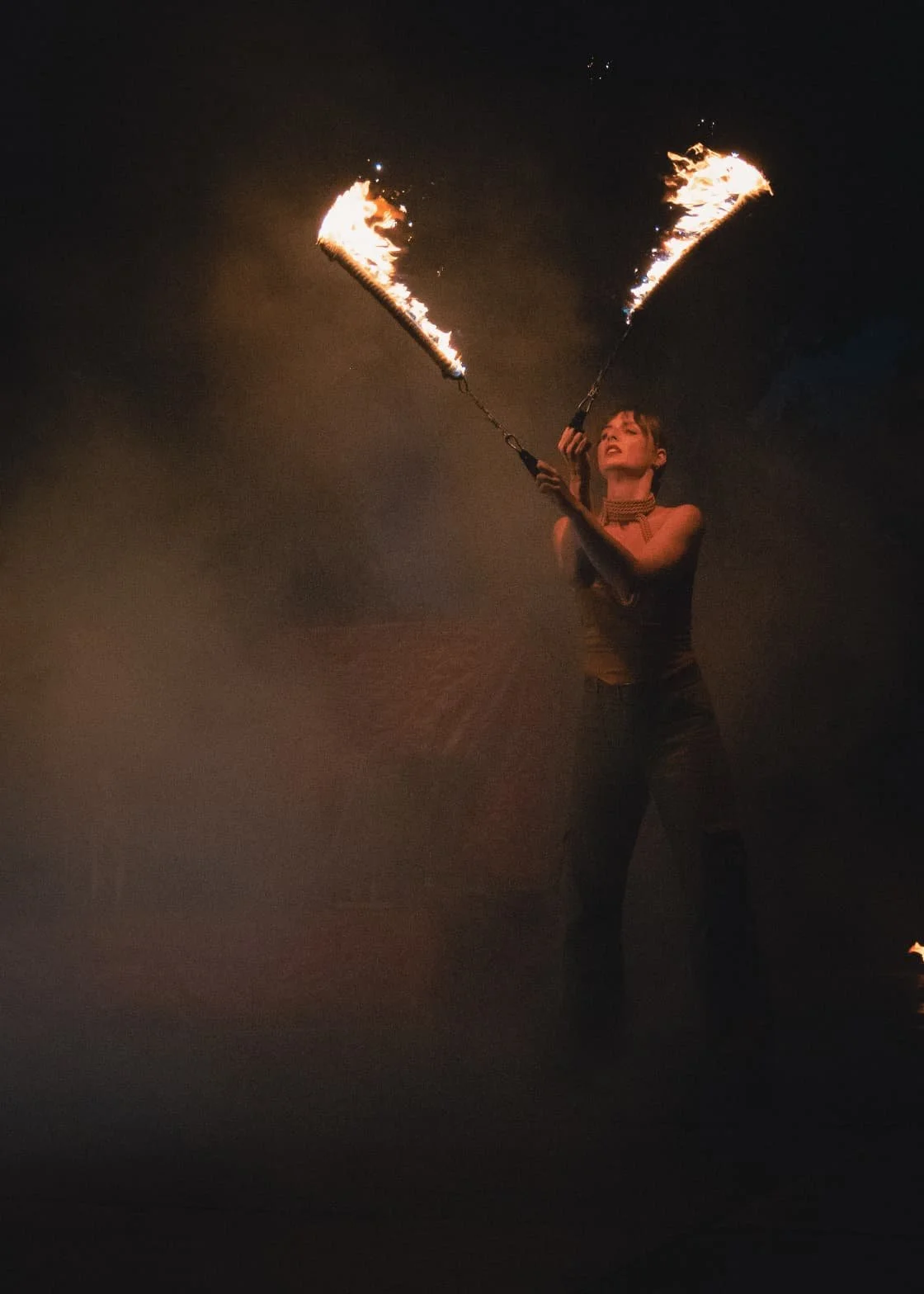 Une femme manipule deux bâtons enflammés lors d'une performance nocturne, le fond est sombre et le feu crée une belle lumière autour d'elle.