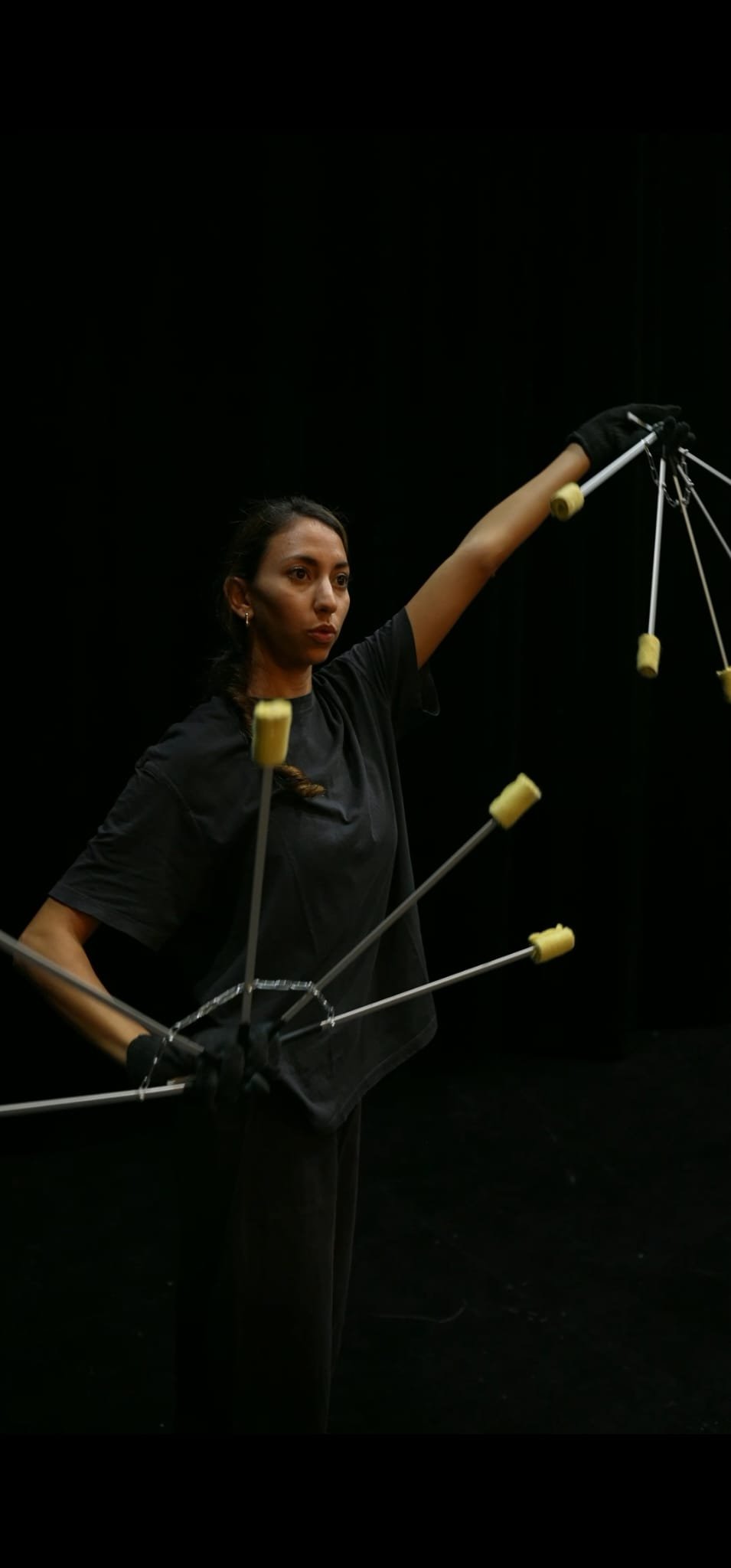 Une femme tient une structure mécanique avec des bras articulés, contre un fond noir, semblable à une scène de théâtre ou une performance artistique.