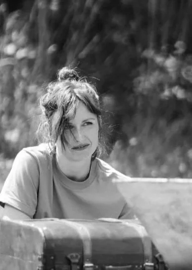 Femme souriante assise à côté d'une valise dehors, probablement lors d'une promenade ou voyage.