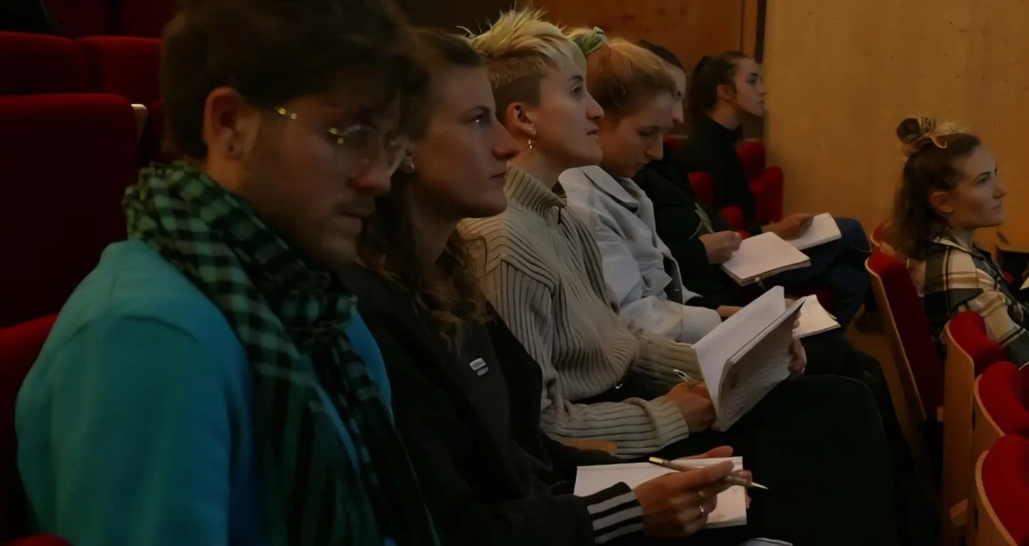 Groupe de jeunes assis dans une salle, certains lisant ou prenant des notes, lors d'une conférence ou d'un atelier.
