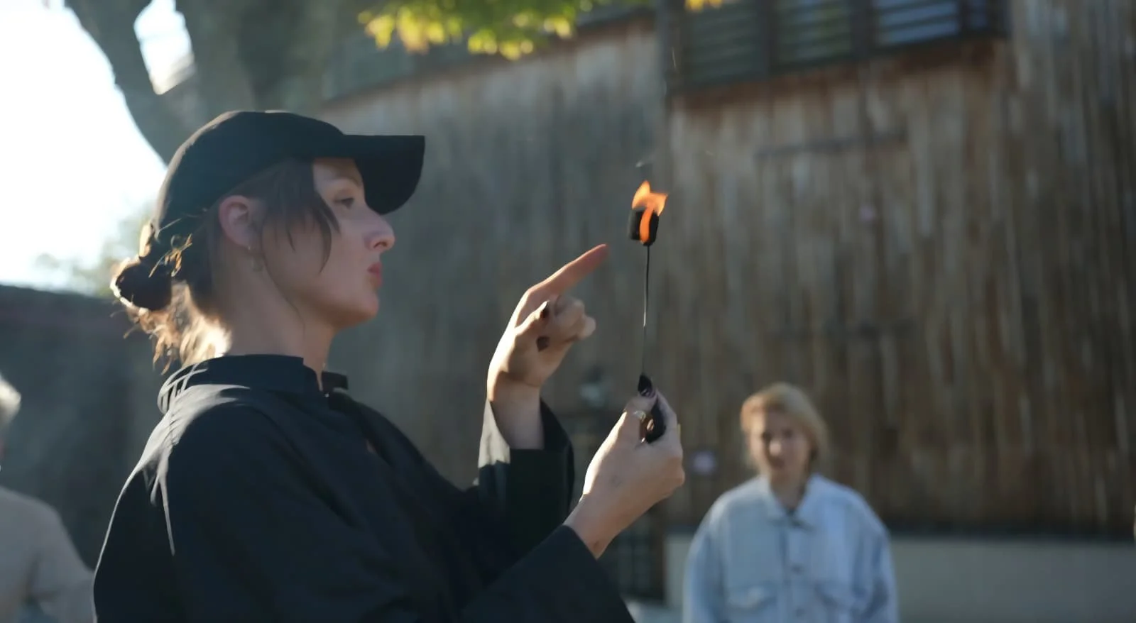 Une femme portant un chapeau noir et une veste noire, tenant une torche enflammée, lors d'une activité en plein air, avec une autre femme en arrière-plan, dans un cadre extérieur avec un mur en bois et des arbres.