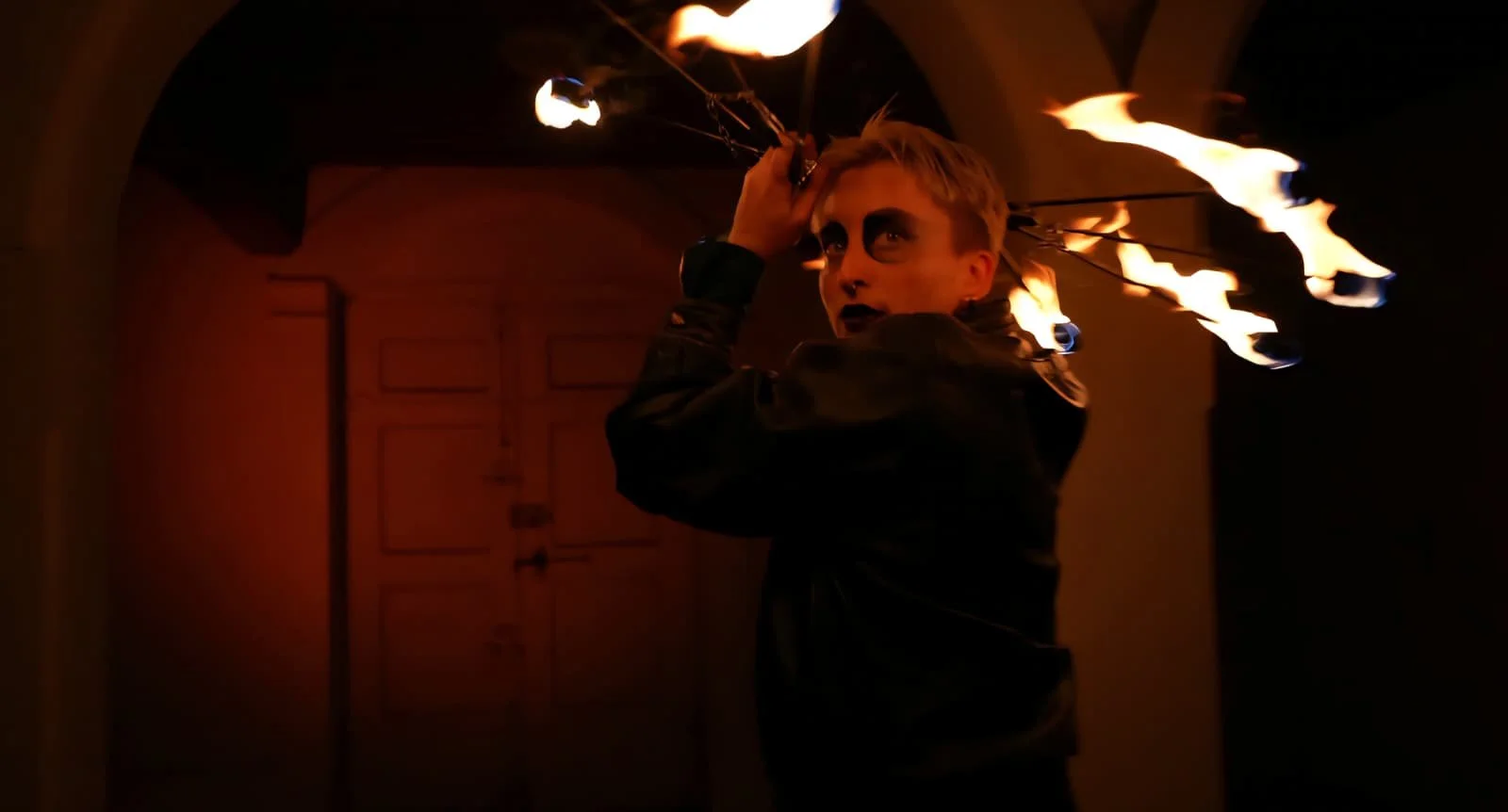 Une personne avec du maquillage noir autour des yeux, manipulant des flambeaux enflammés dans une pièce sombre.