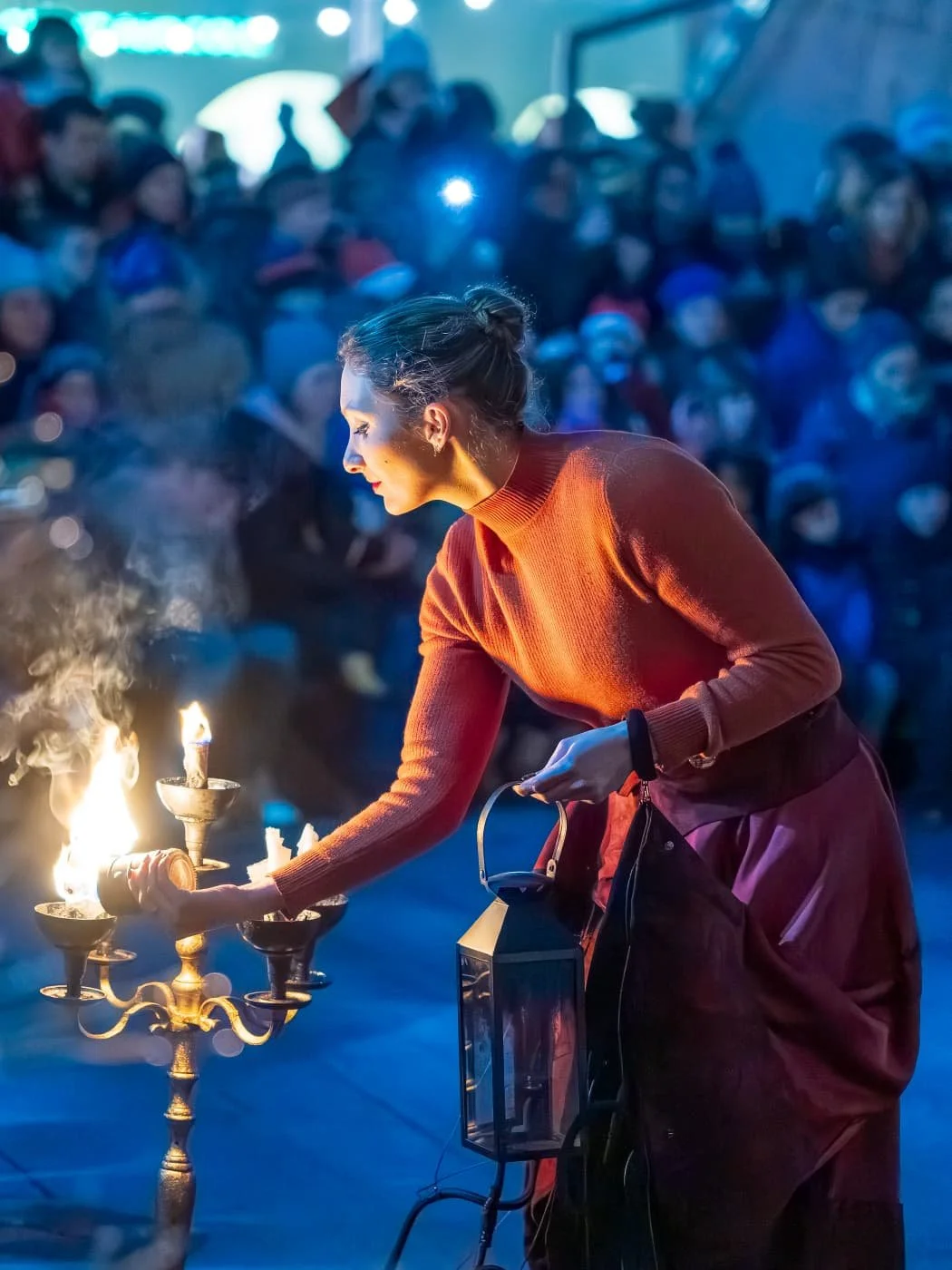 Une femme dépose une bougie allumée sur une chandelier en laiton lors d'une cérémonie ou d'un rituel, tandis qu'un grand groupe de personnes regarde en arrière-plan.