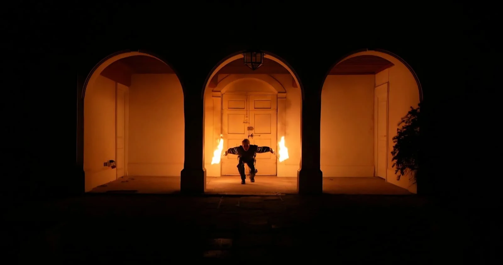 Une personne en costume noir réalise un spectacle de feu lors d'une performance nocturne dans un espace architectural avec trois arches, le centre étant illuminé par des flammes.