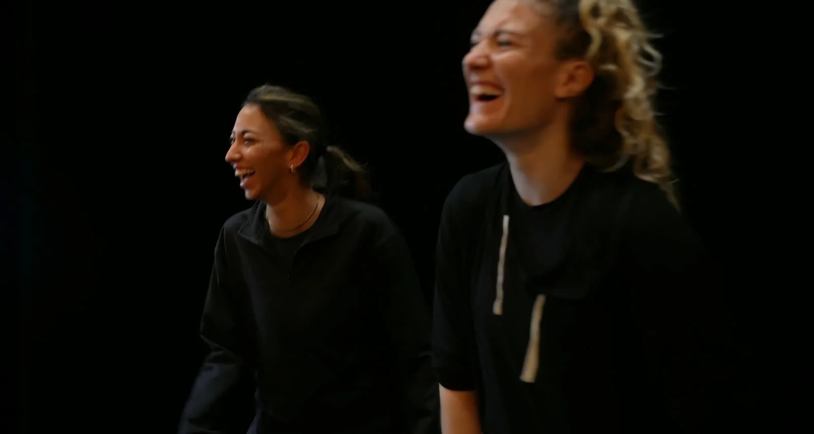 Deux femmes rient et sourient dans un environnement noir, portant des vêtements noirs.