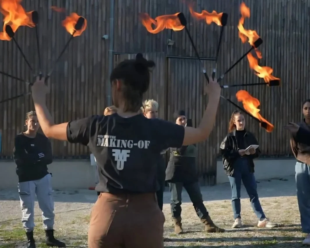 Une femme effectue un jonglage avec des torches enflammées lors d'une performance en extérieur, plusieurs spectateurs regardent.