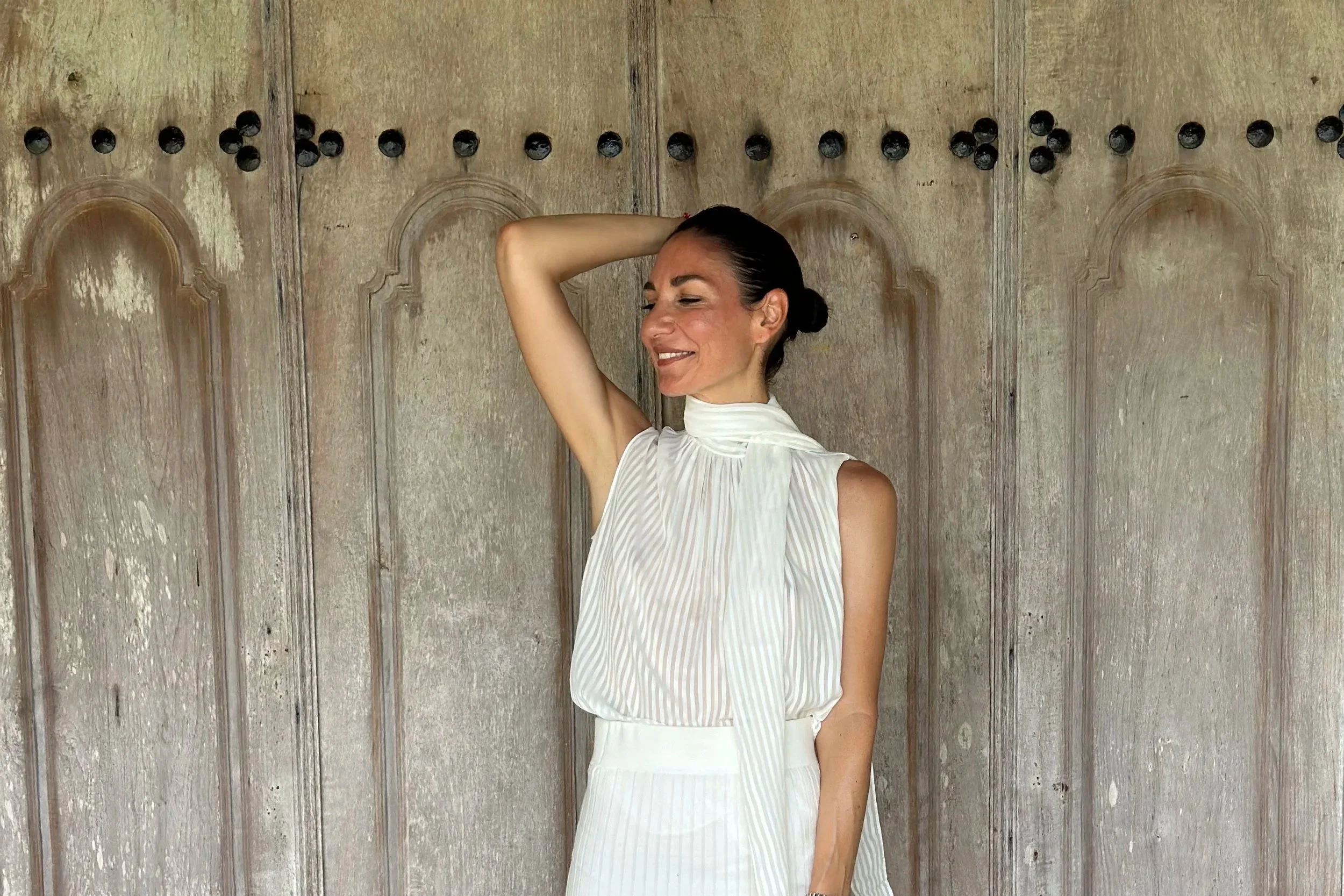 Smiling woman, peaceful demeanour, with a white dress, in front of a wooden door.
Femme souriante, visage paisible, avec une robe blanche, posant devant une porte en bois.