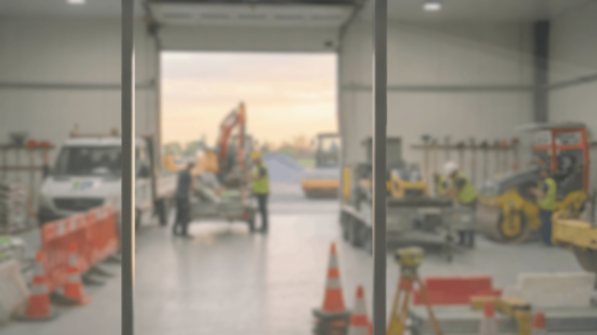 Photo floue d'un espace de construction ou de rénovation avec des travailleurs en vestes fluorescentes, des équipements et des véhicules, vus à travers une structure en métal verticale.