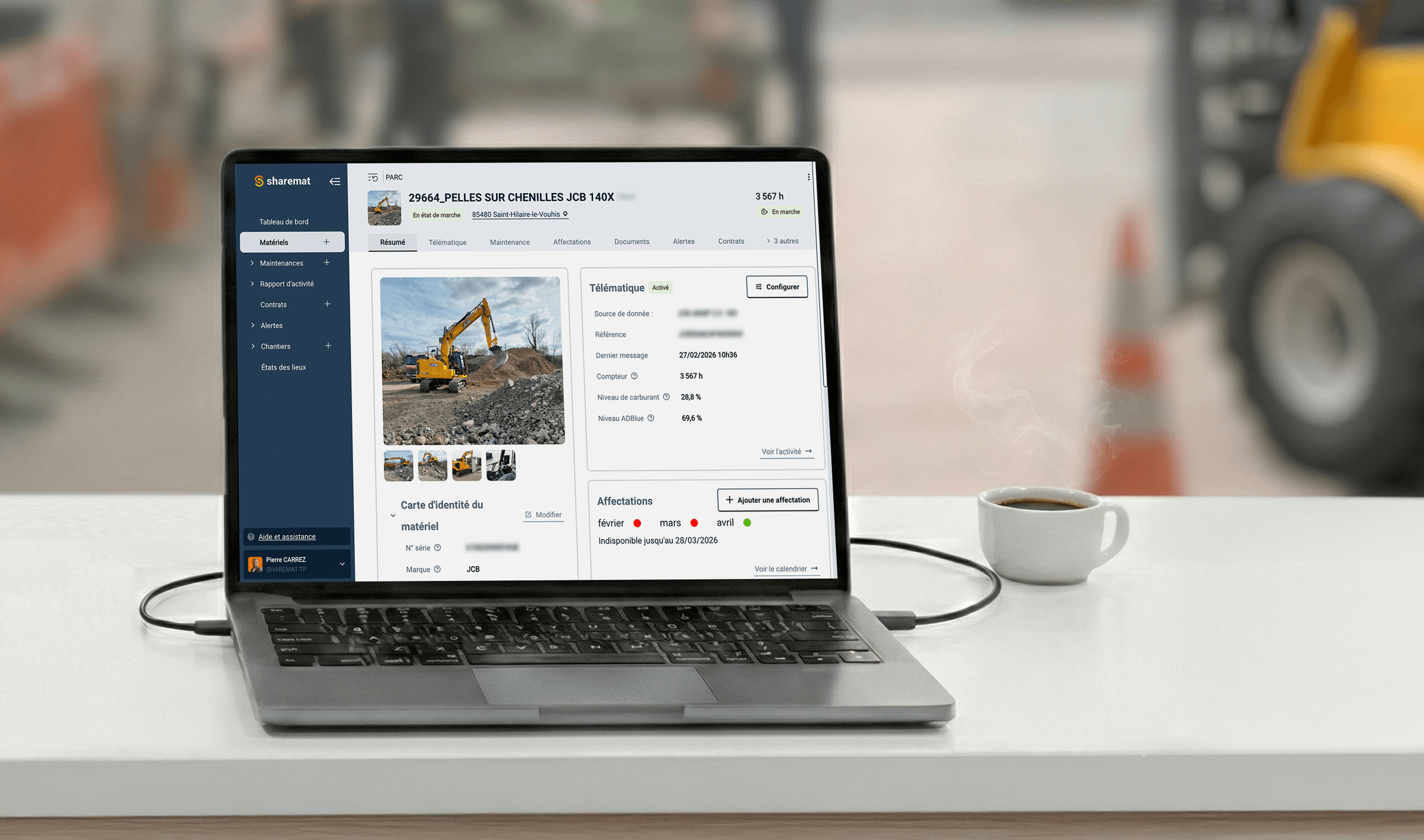 Un ordinateur portable affichant une plateforme de gestion de matériel de construction avec une image de pelle sur un chantier, sur un bureau avec une tasse de café à côté.