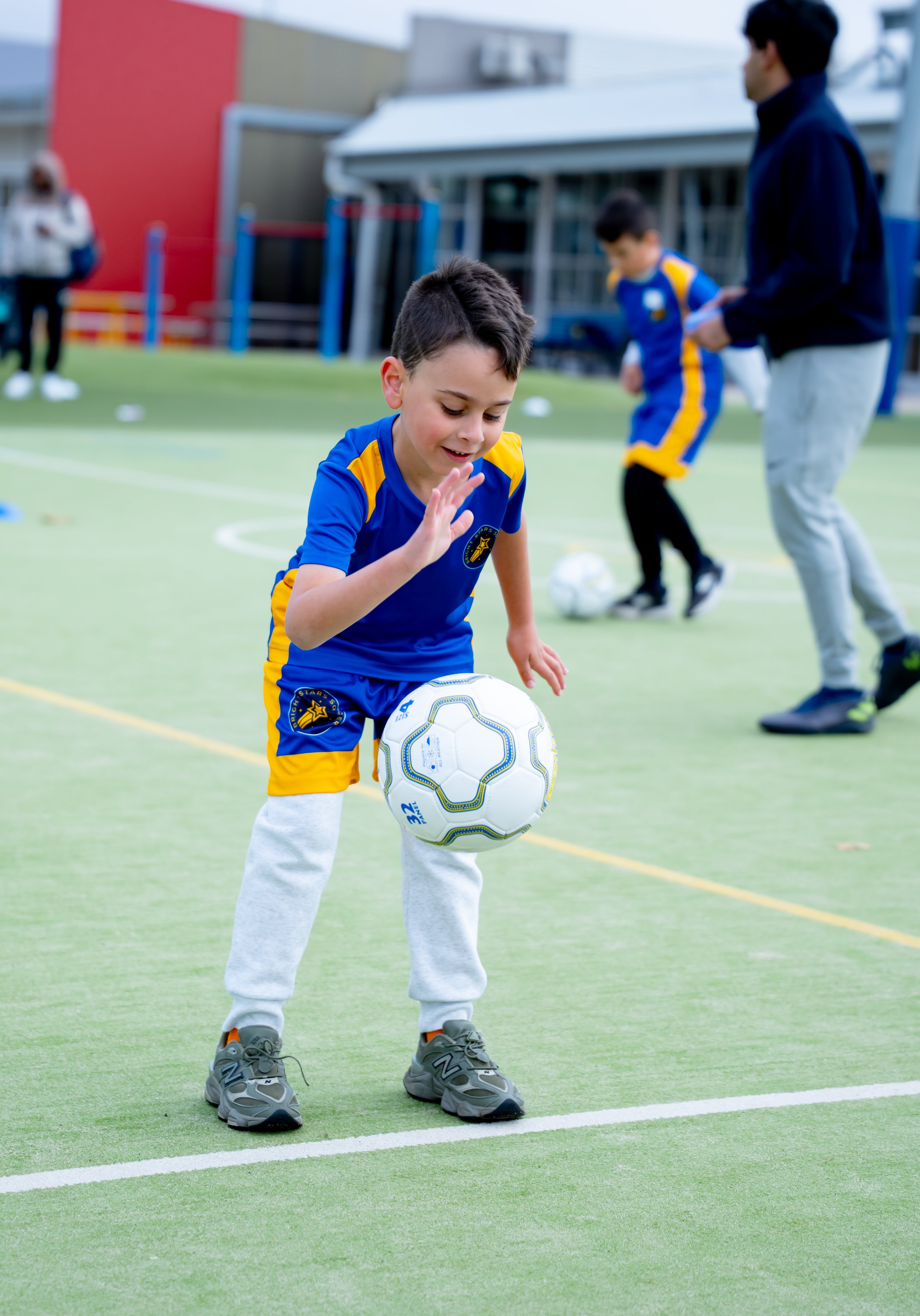 Young boy wearing a blue sports uniform with yellow accents, standing on a sports field, balancing a soccer ball on his feet.