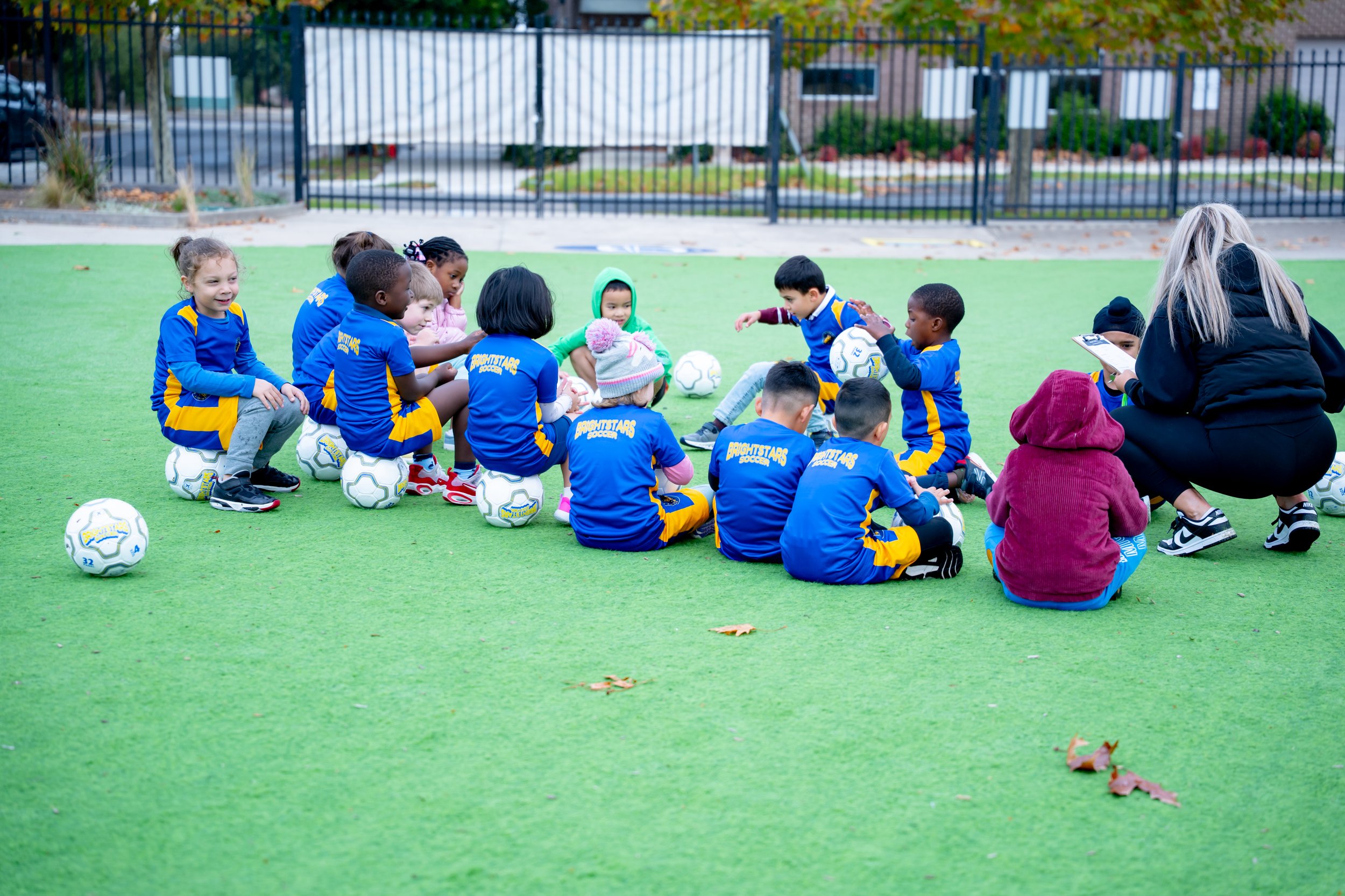 Children in blue and yellow soccer uniforms sitting on a green turf field, listening to a Bright Stars Soccer Coach with a clipboard