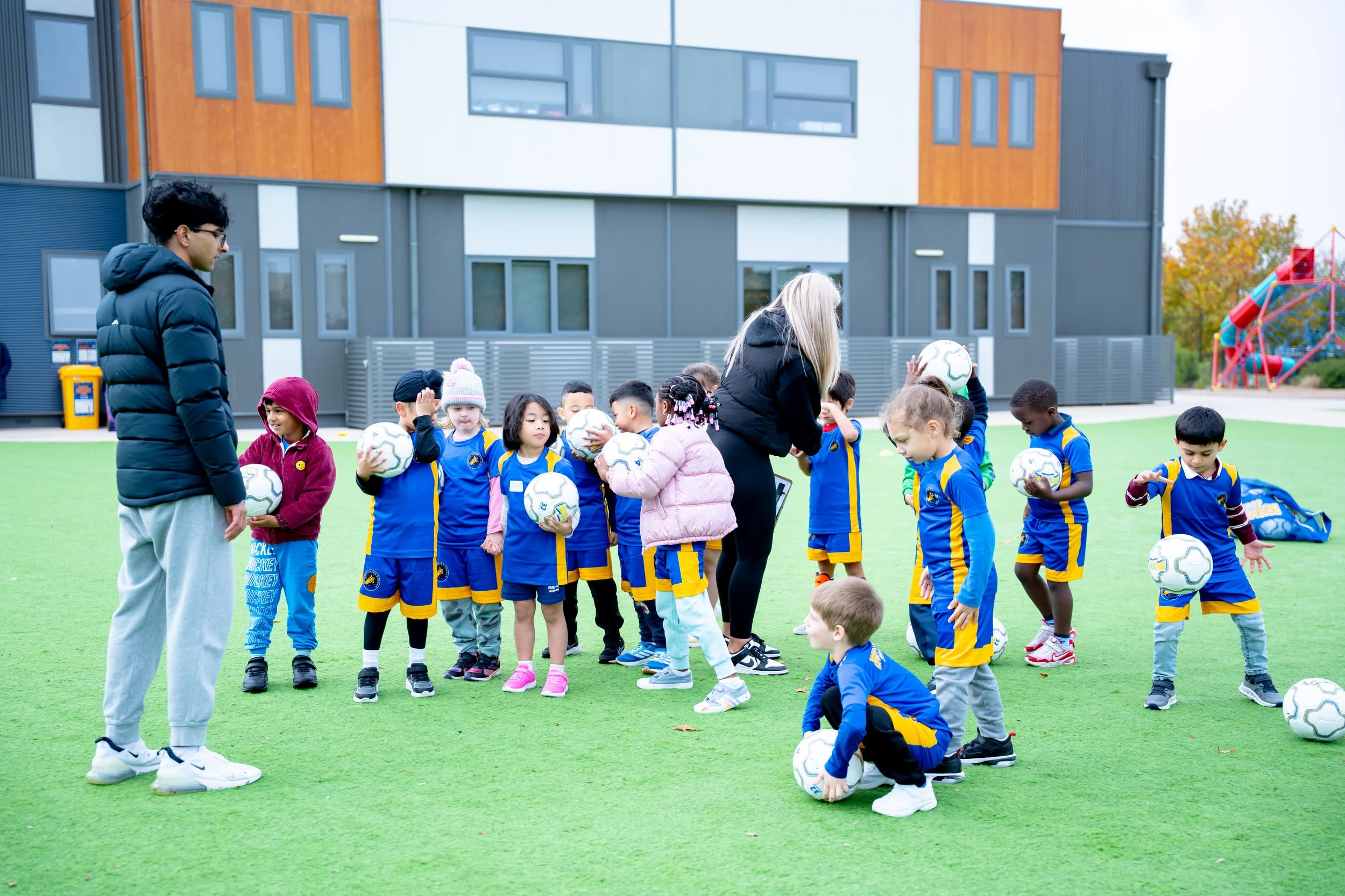 Group of young children in blue and yellow sports uniforms, holding soccer balls during outdoor soccer practice 