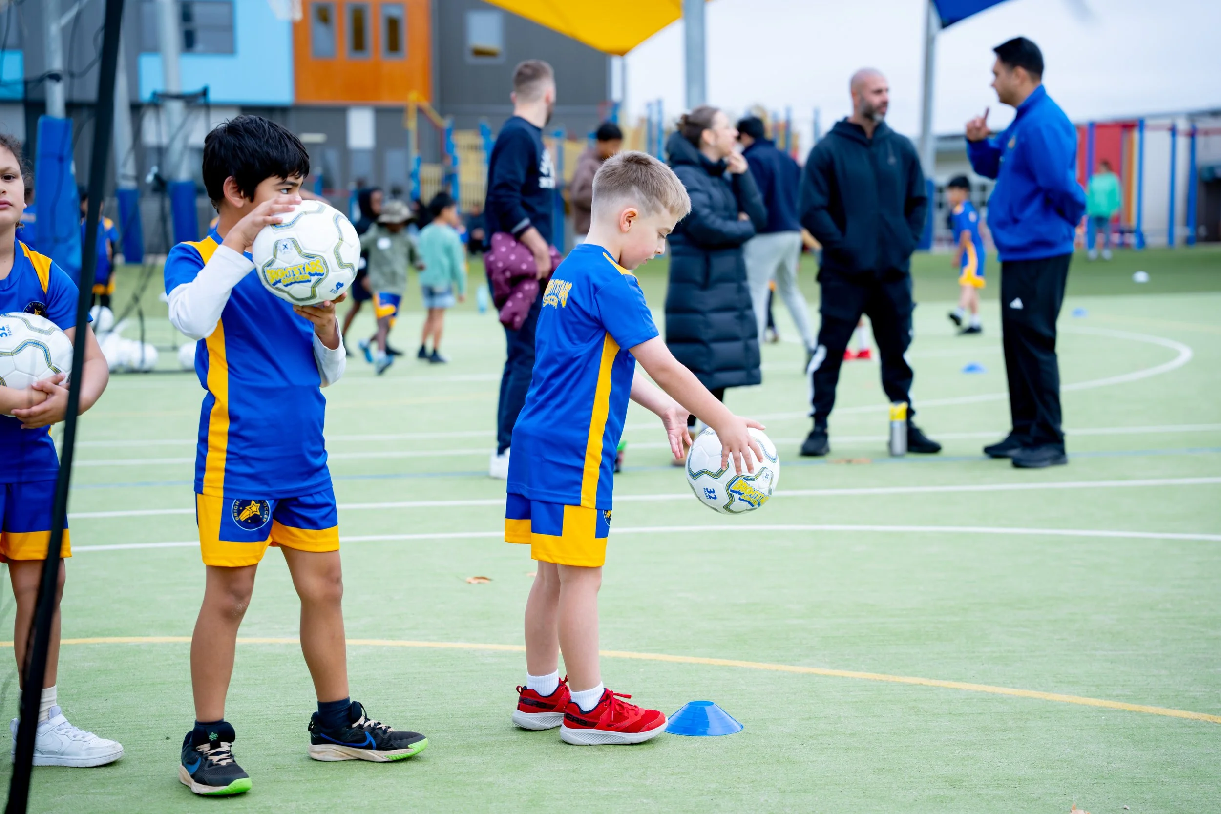 Young children in blue sports uniforms participating in a soccer practice or game on an outdoor field, with coaches and spectators watching.