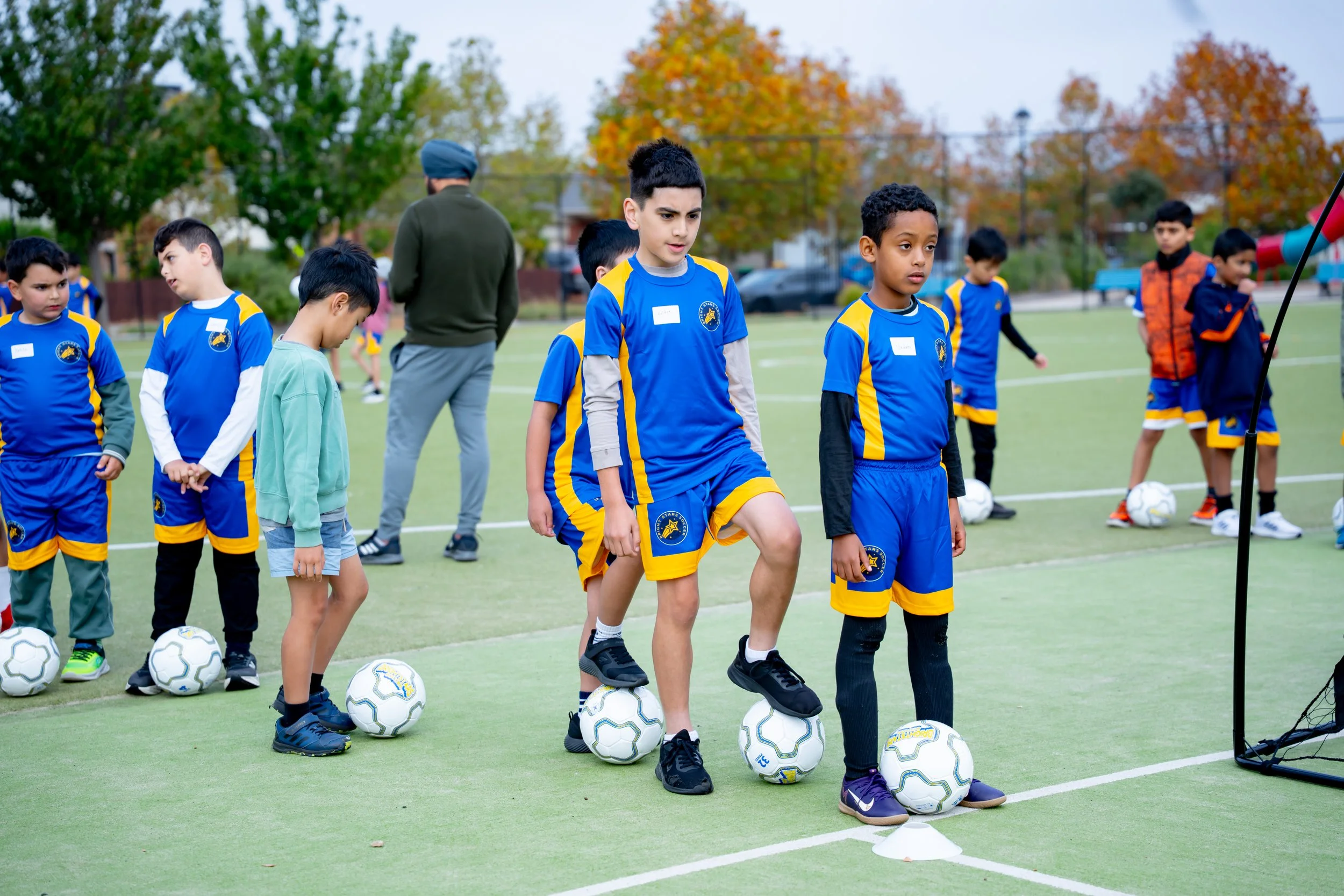 Group of young boys wearing blue and yellow Bright Stars Soccer uniforms, standing in a line on a soccer field, each with a soccer ball at their feet, preparing for soccer practice