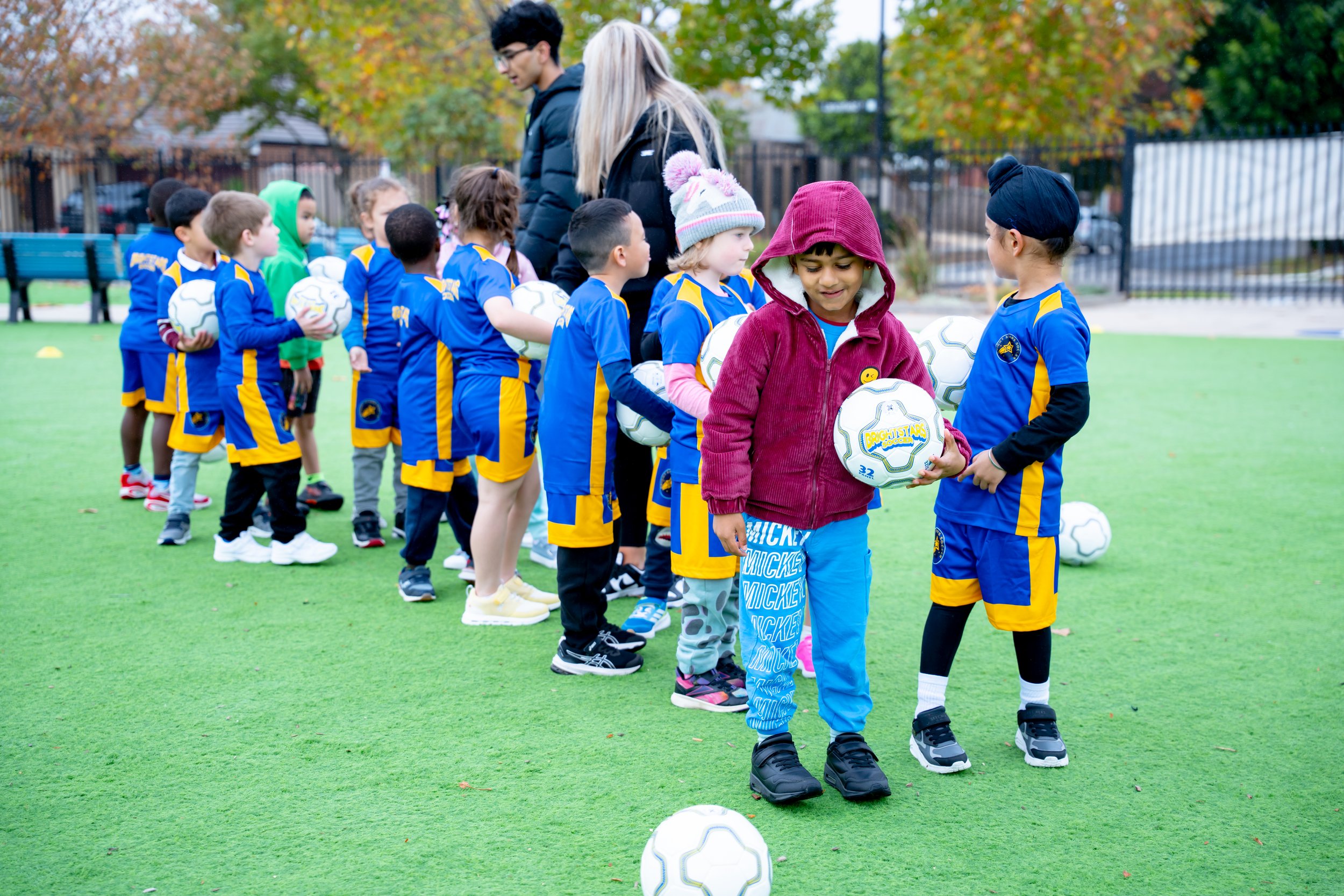 Children in blue and yellow soccer uniforms on a field, lined up with balls, listening to Bright Stars Soccer coach