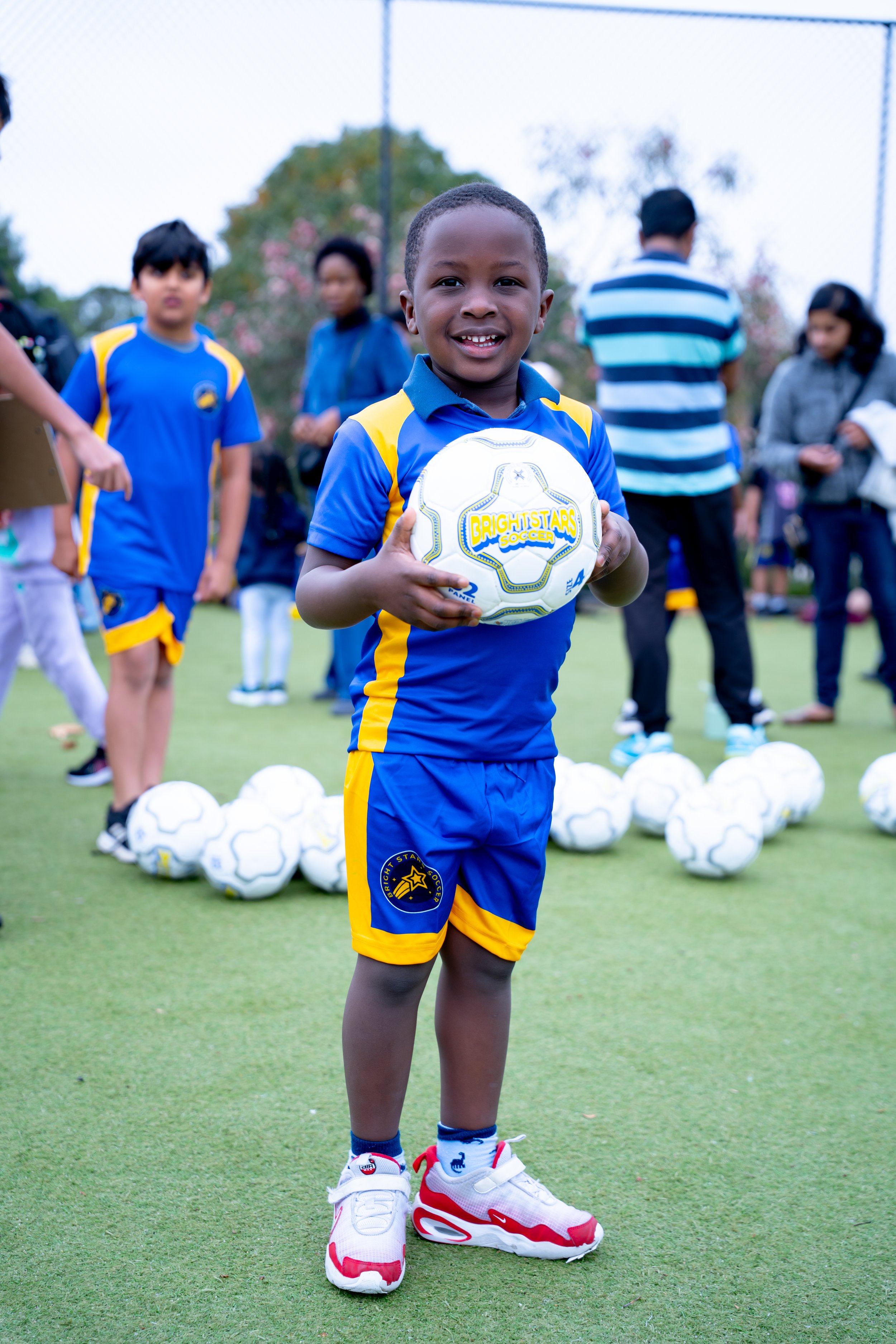 A young boy in a blue and yellow Bright Stars Soccer uniform holding a soccer ball, standing on a grass field surrounded by children and adults.