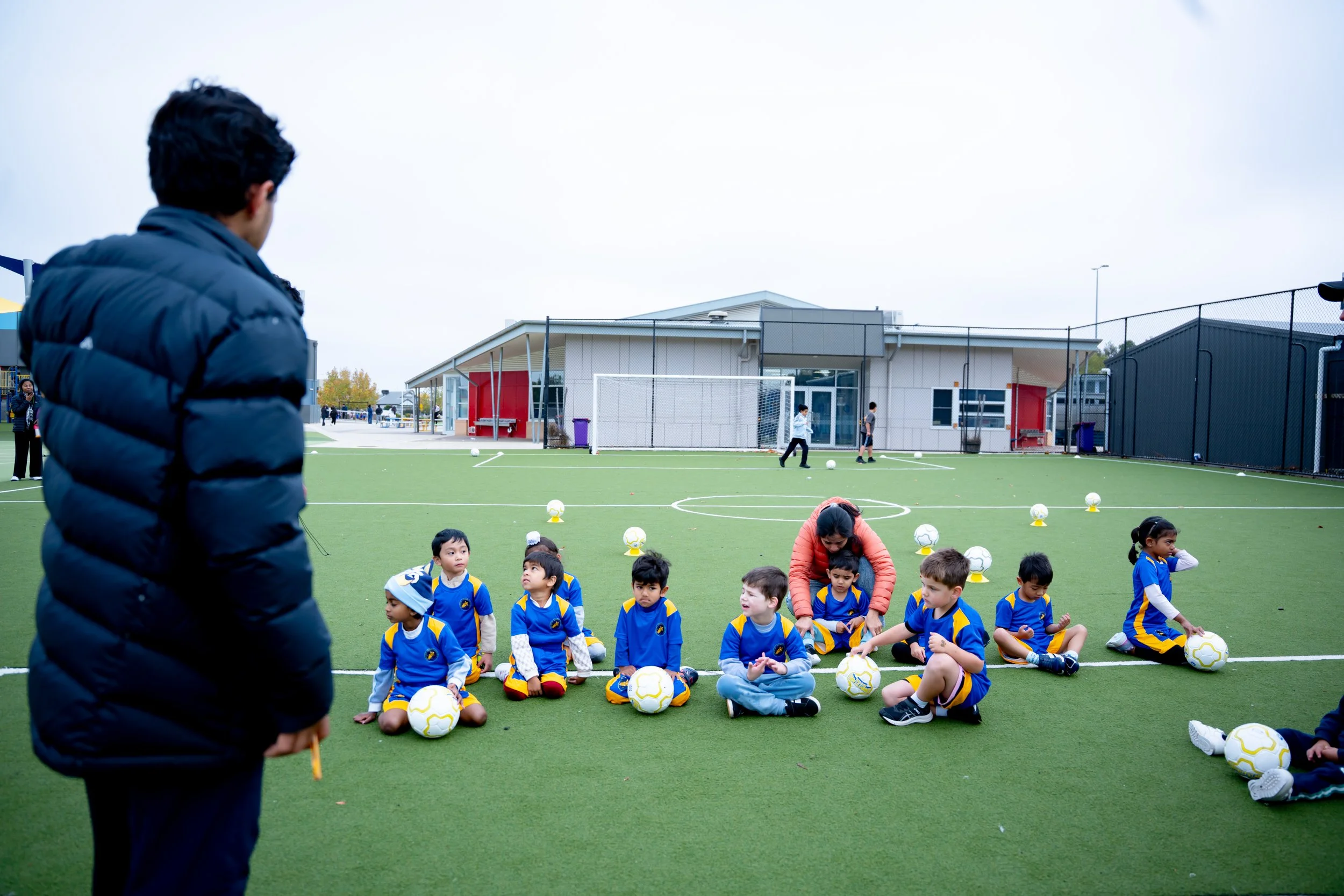 A group of young children in Bright Stars Soccer uniforms with soccer balls, sitting on a green artificial turf field during a soccer practice 