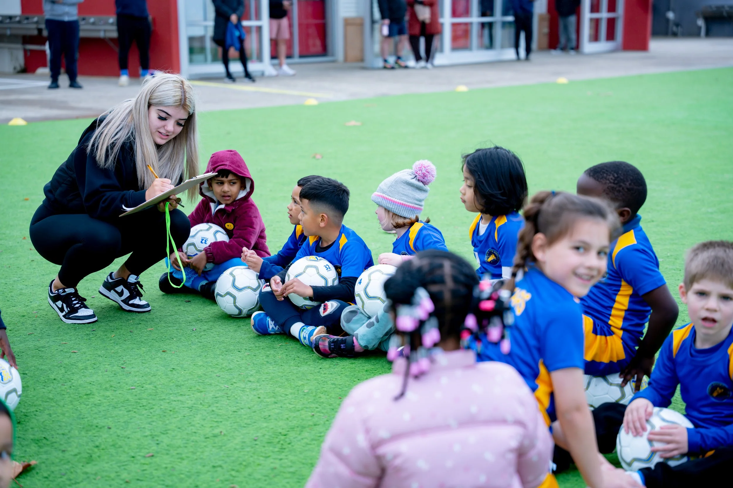 A group of young children in blue and yellow sports uniforms sitting on a green field with soccer balls