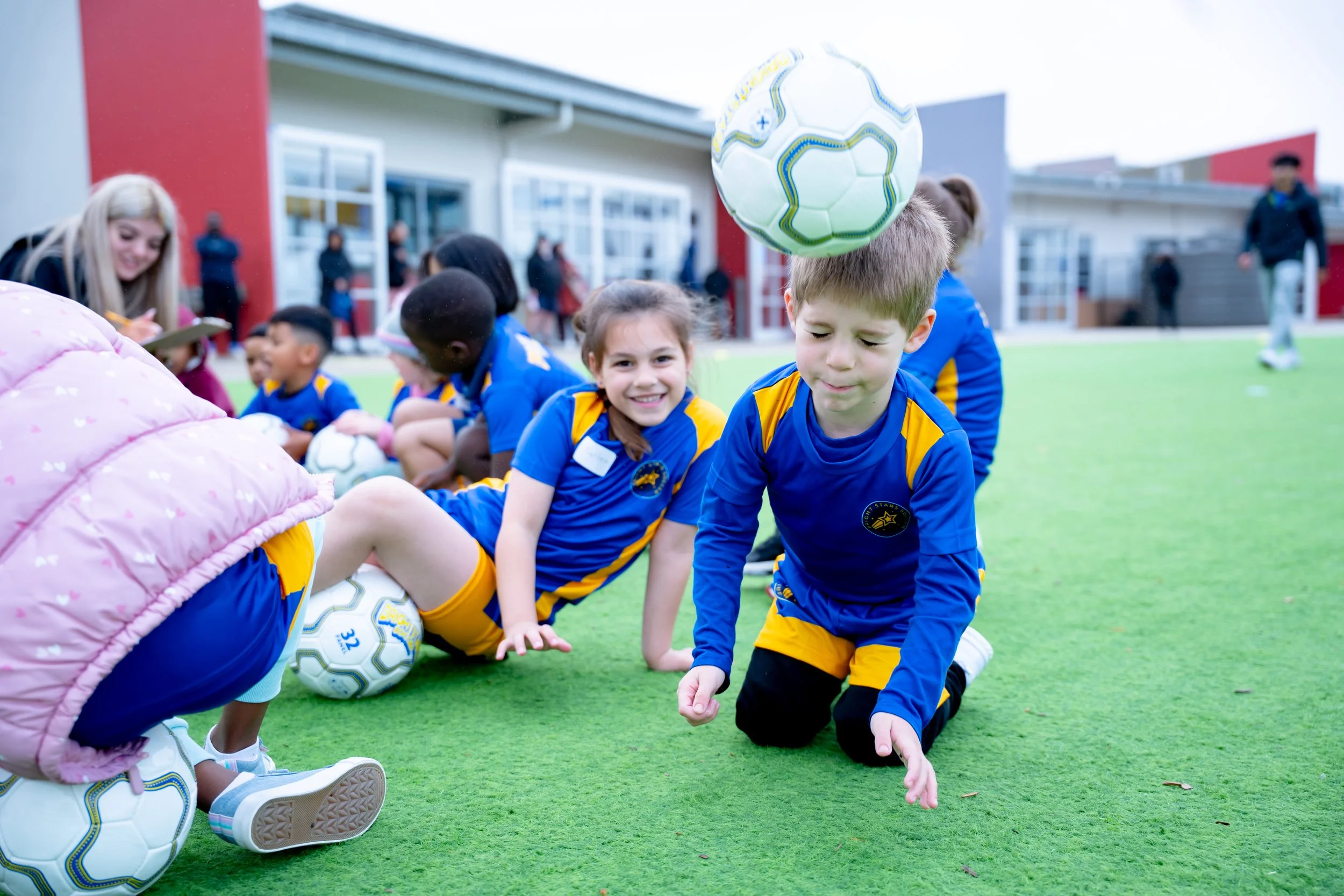 Kids in Bright Stars Soccer uniforms sitting on artificial turf, with a soccer ball