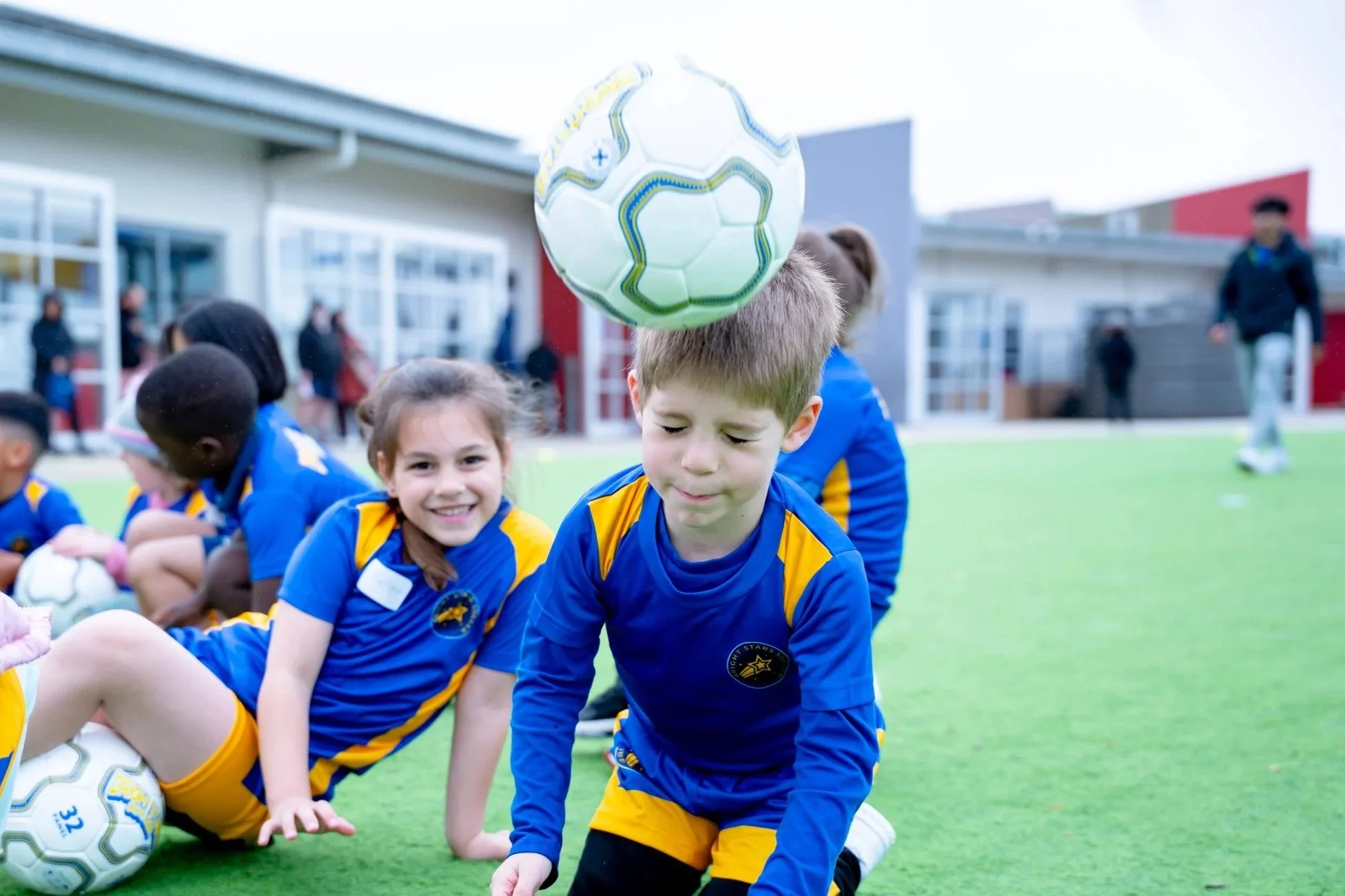 Young children in Bright Stars Soccer uniforms sitting on the grass during a team event, with soccer balls nearby.
