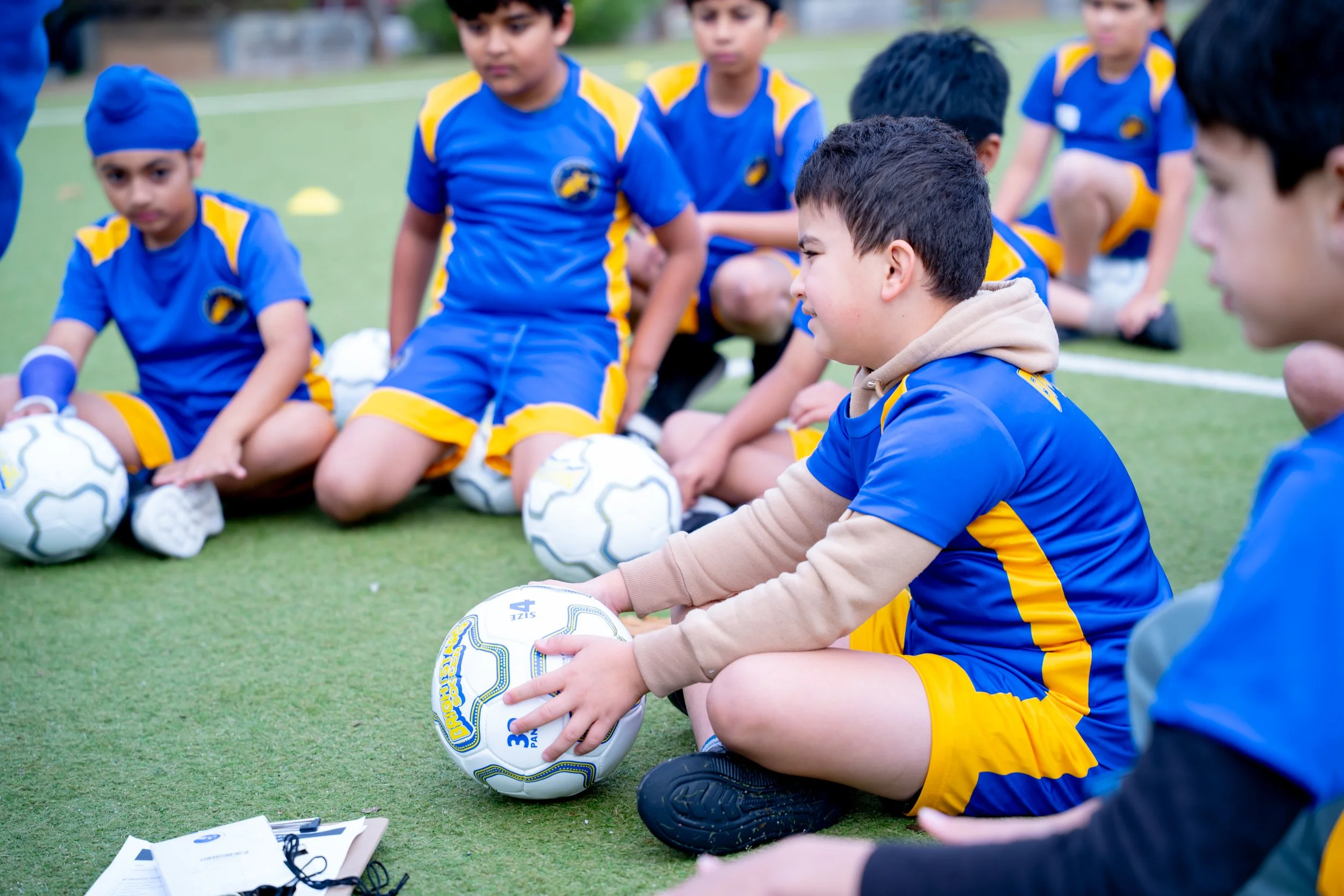 Group of young boys in blue and yellow sports uniforms sitting on a grassy field during a soccer practice