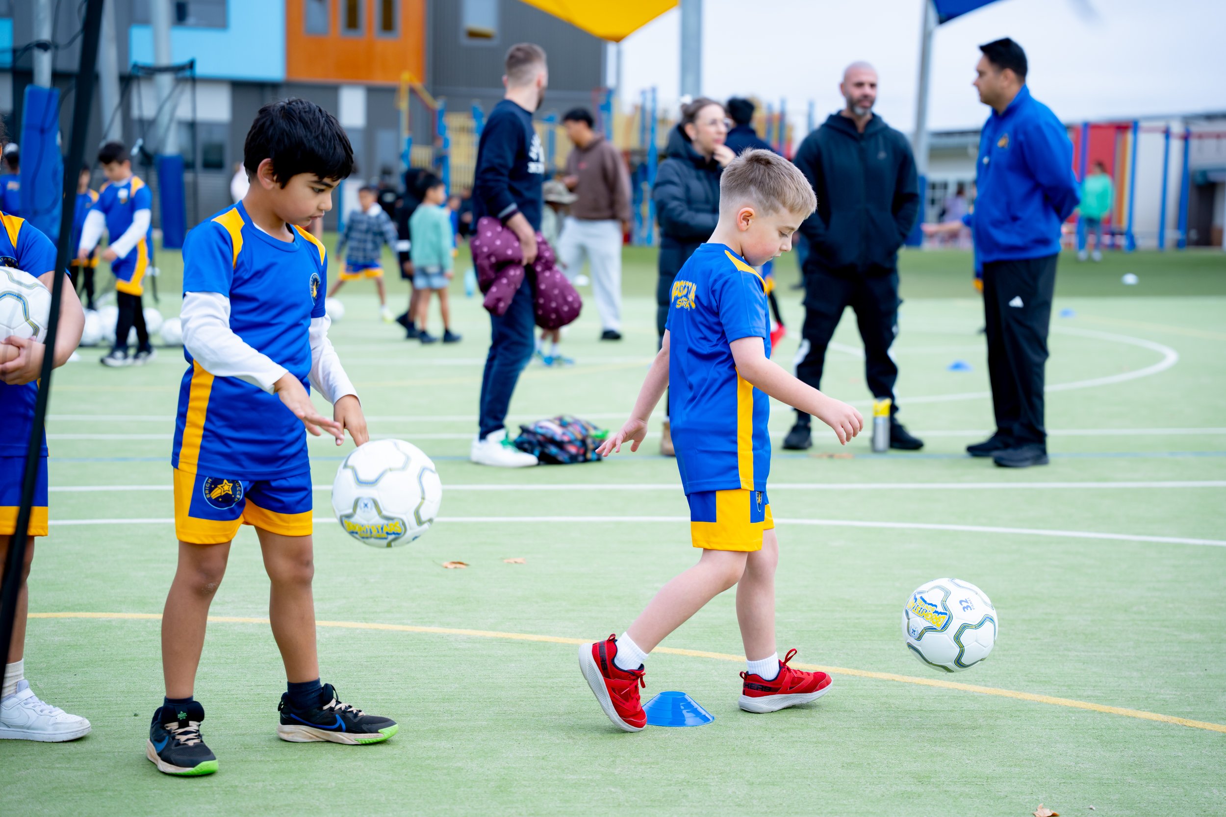 Young children in blue and yellow sports uniforms practicing soccer on an outdoor field with adults and other children in the background.