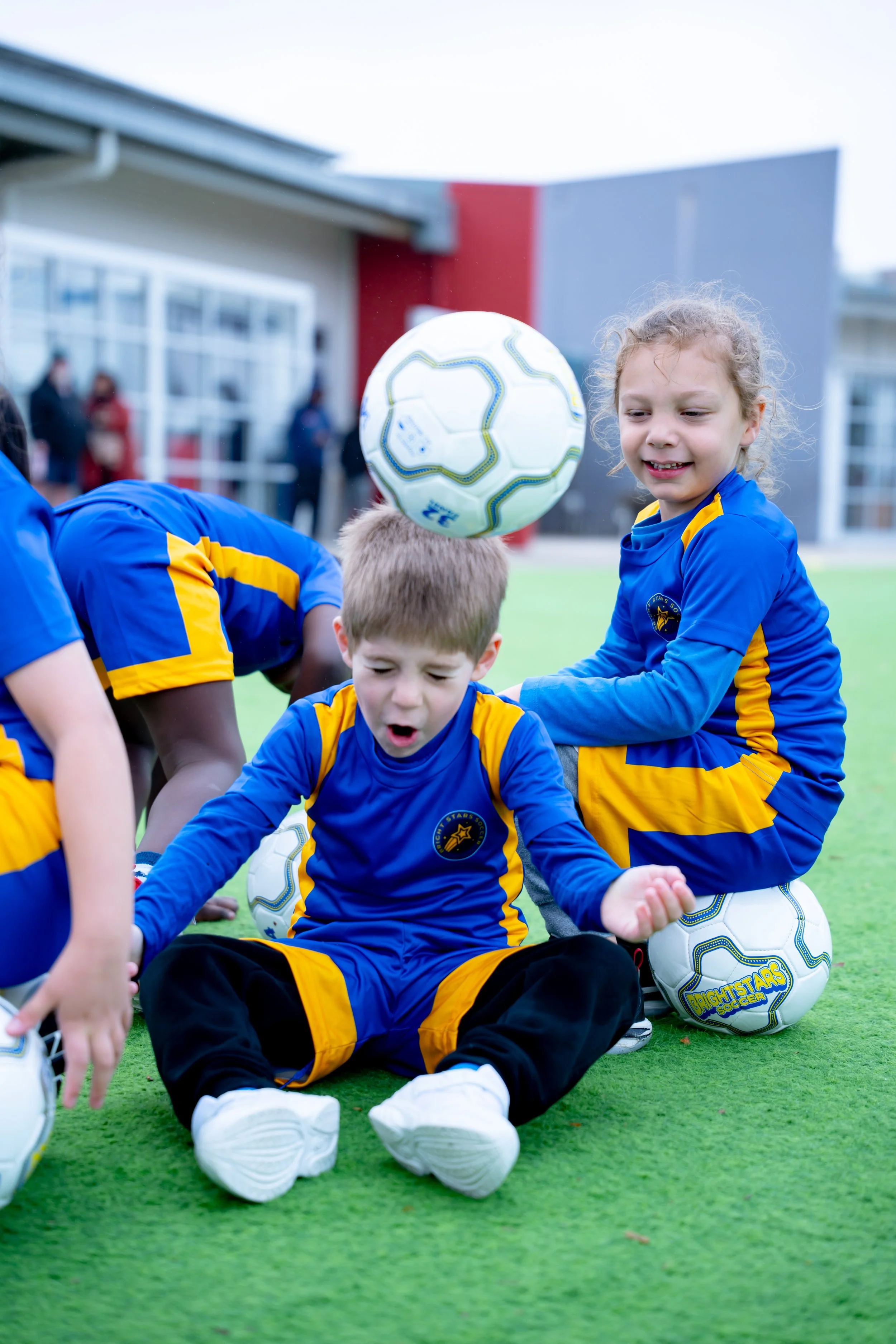 Young children wearing Bright Stars Soccer uniforms sitting on the grass, with some children controlling and balancing soccer balls during a soccer practice or game.