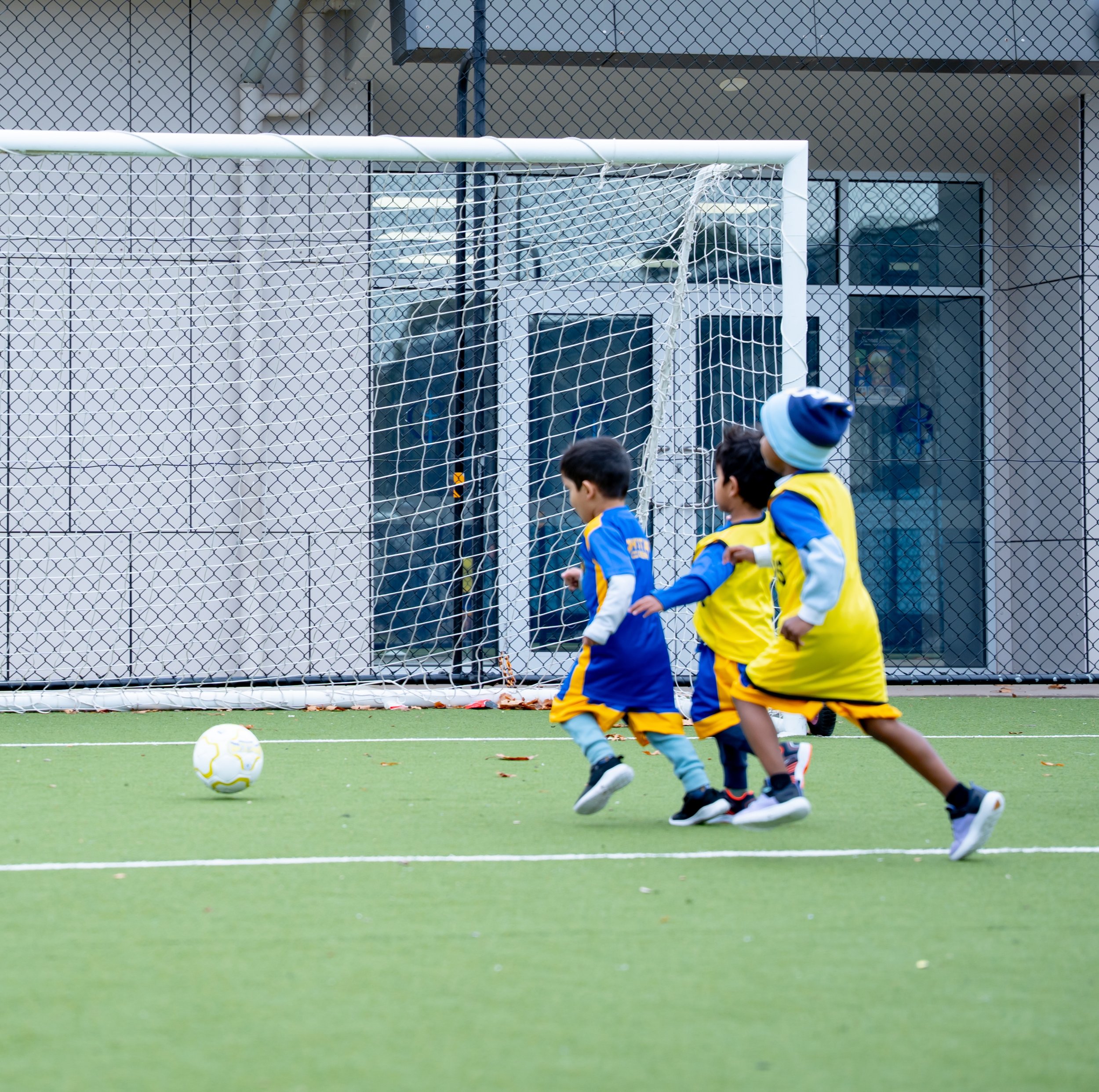 A group of children playing soccer on a green field, with a goal post and fence in the background.