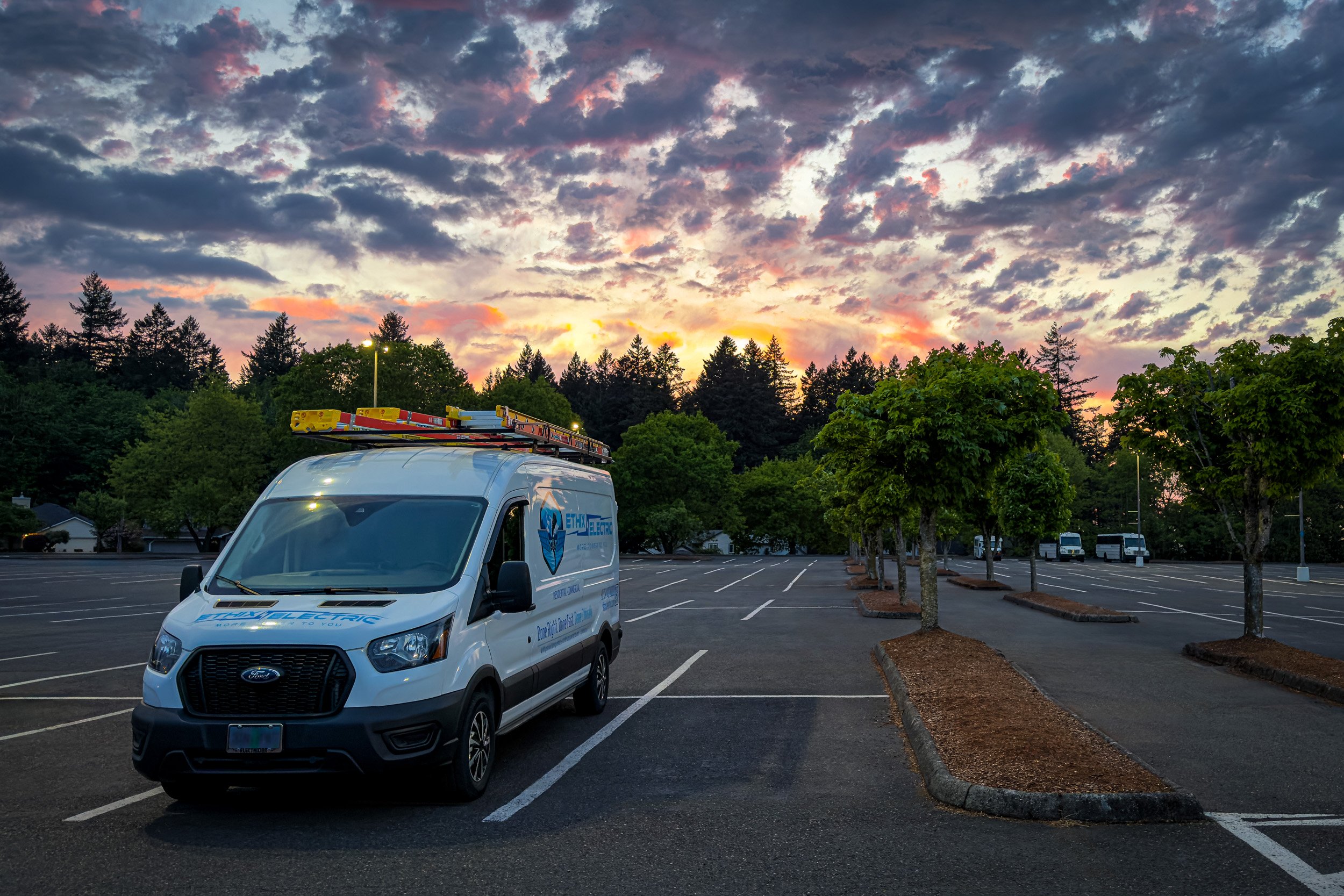 EthiX Electric service van under a dramatic Portland evening sky at sunset, with red and yellow horizon colors, cool blue tones above, and green trees surrounding a parking lot.