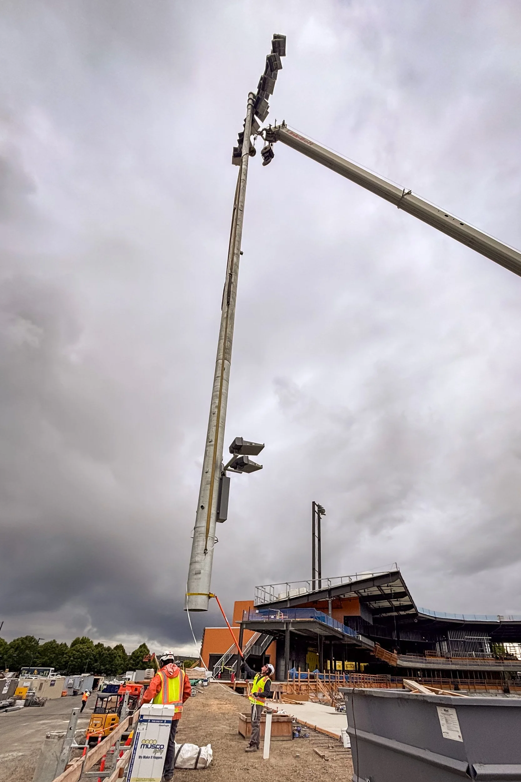EthiX Electric founder in safety vest and PPE guiding a giant stadium light pole from the bottom while a crane lifts it at the Hillsboro Hops Stadium construction site.