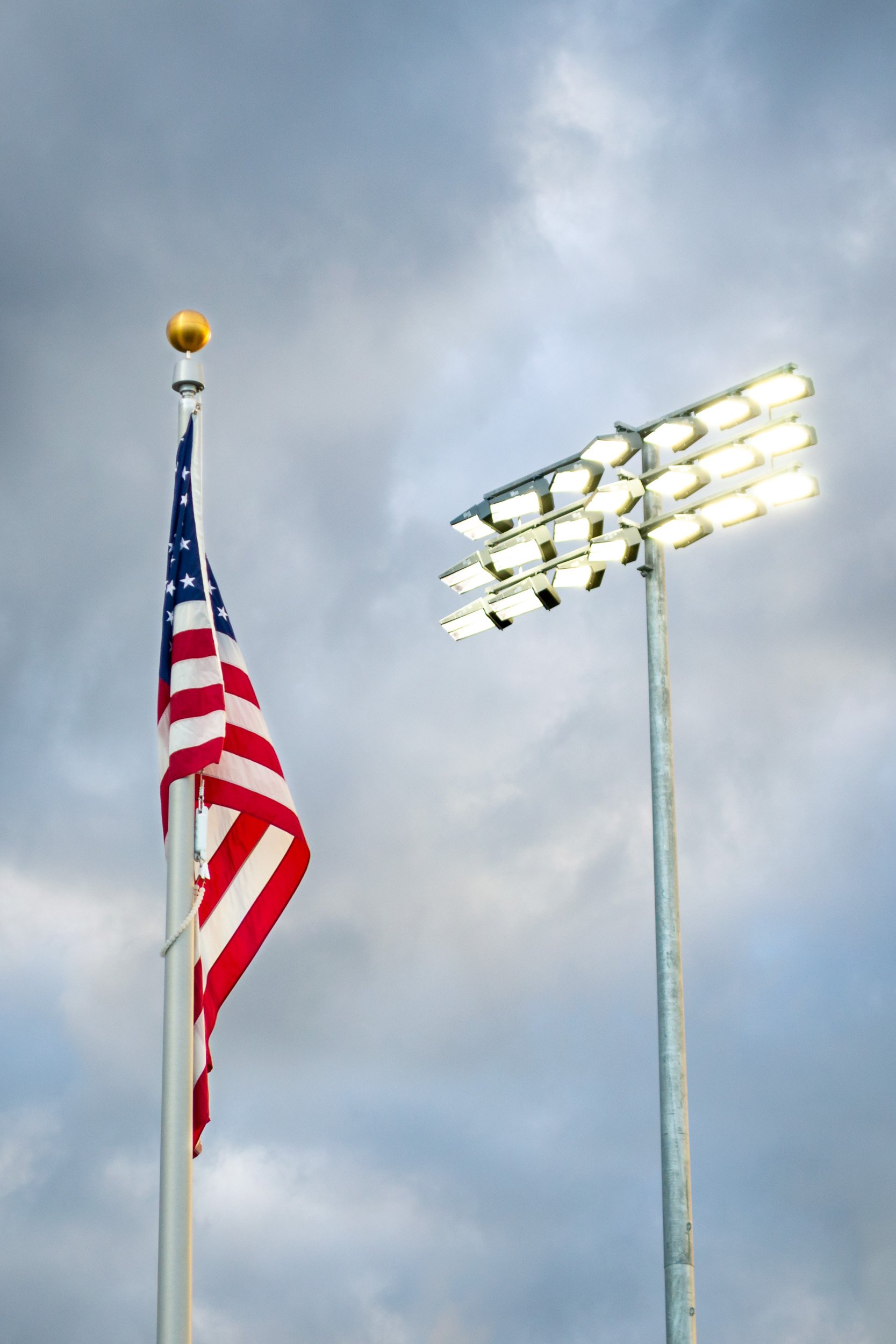 Completed Hillsboro Hops stadium lighting shining toward a large flying American flag under a dramatic Hillsboro Oregon sky.