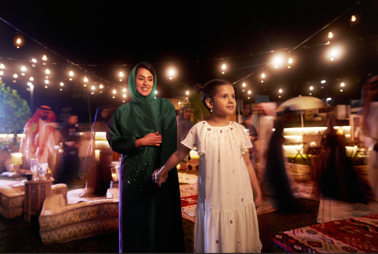 A woman and a young girl holding hands at an outdoor evening event with string lights overhead. The woman is wearing a dark green traditional dress with a hood, and the girl is in a white dress with embroidered details. There are blurred figures and 