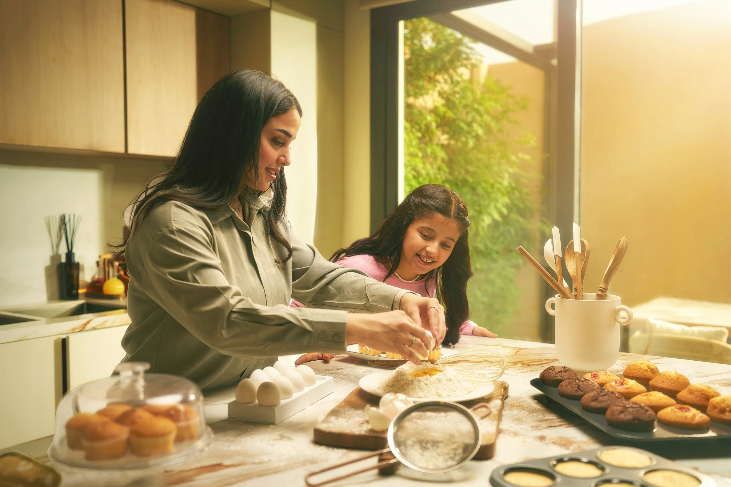 A woman and a girl baking cookies in a kitchen with sunlight streaming in, surrounded by baking ingredients and utensils.