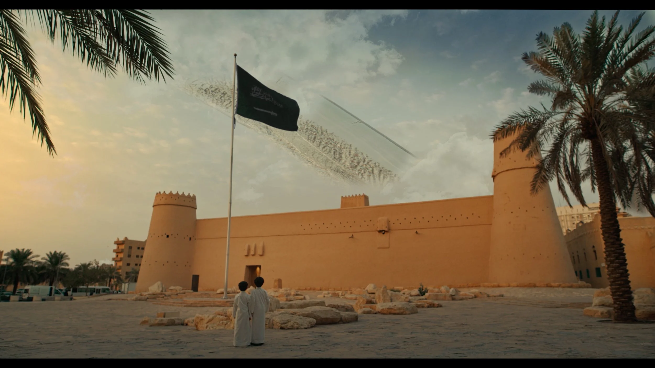 A historical fortress in an outdoor setting with palm trees, a large black flag, and two people in traditional white attire standing in front of the fortress, during a clear day.