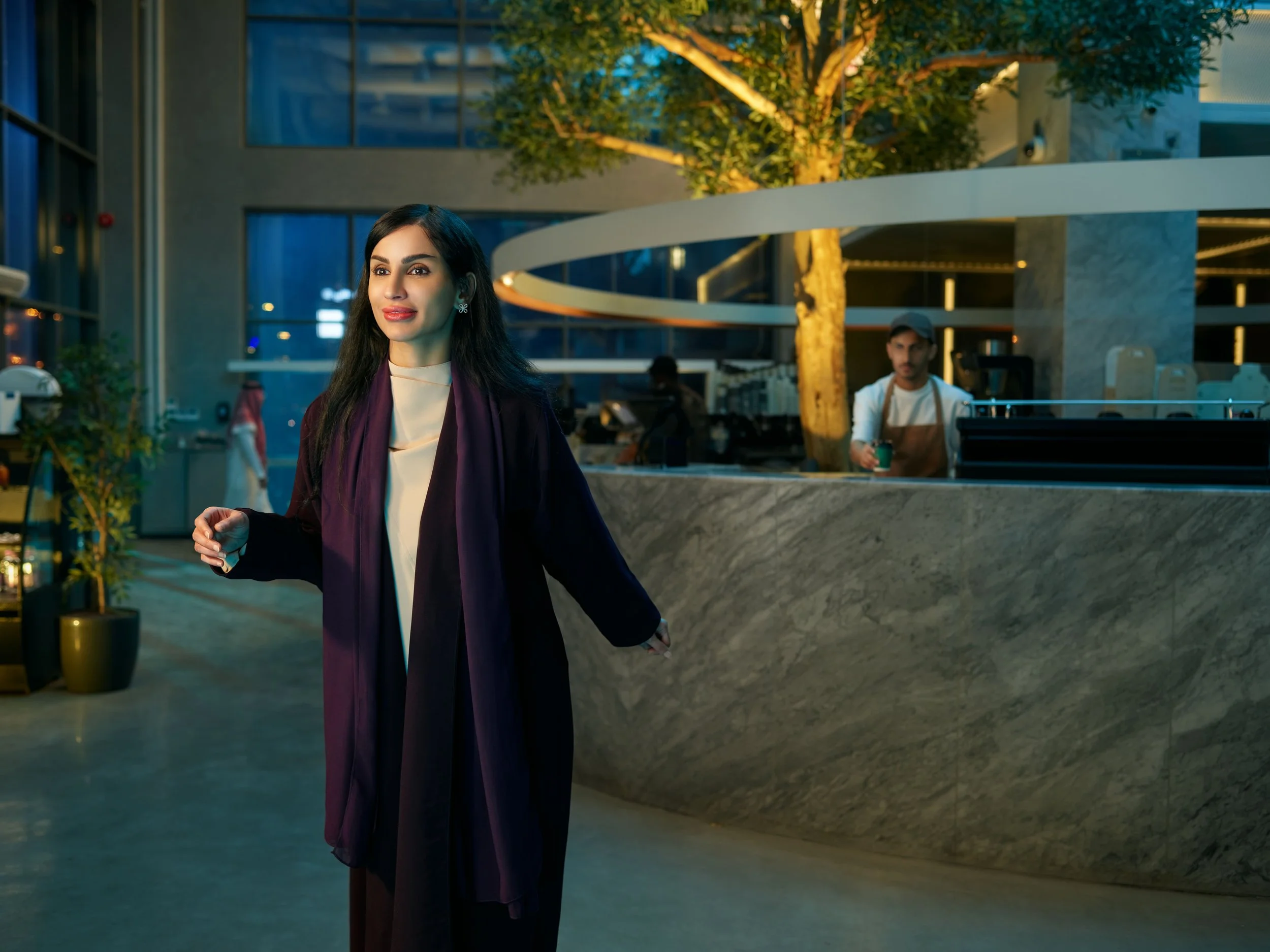 A woman in a formal black and white outfit with a purple scarf walking through a modern indoor space, possibly a café or airport lounge, with a man working behind a marble counter in the background.