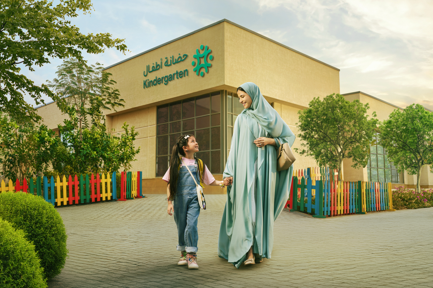 A woman in a hijab and traditional dress holding hands with a young girl outside a kindergarten building. The building has a sign with the word 'Kindergarten' in English and Arabic. There are colorful fences, trees, and a clear sky in the background.