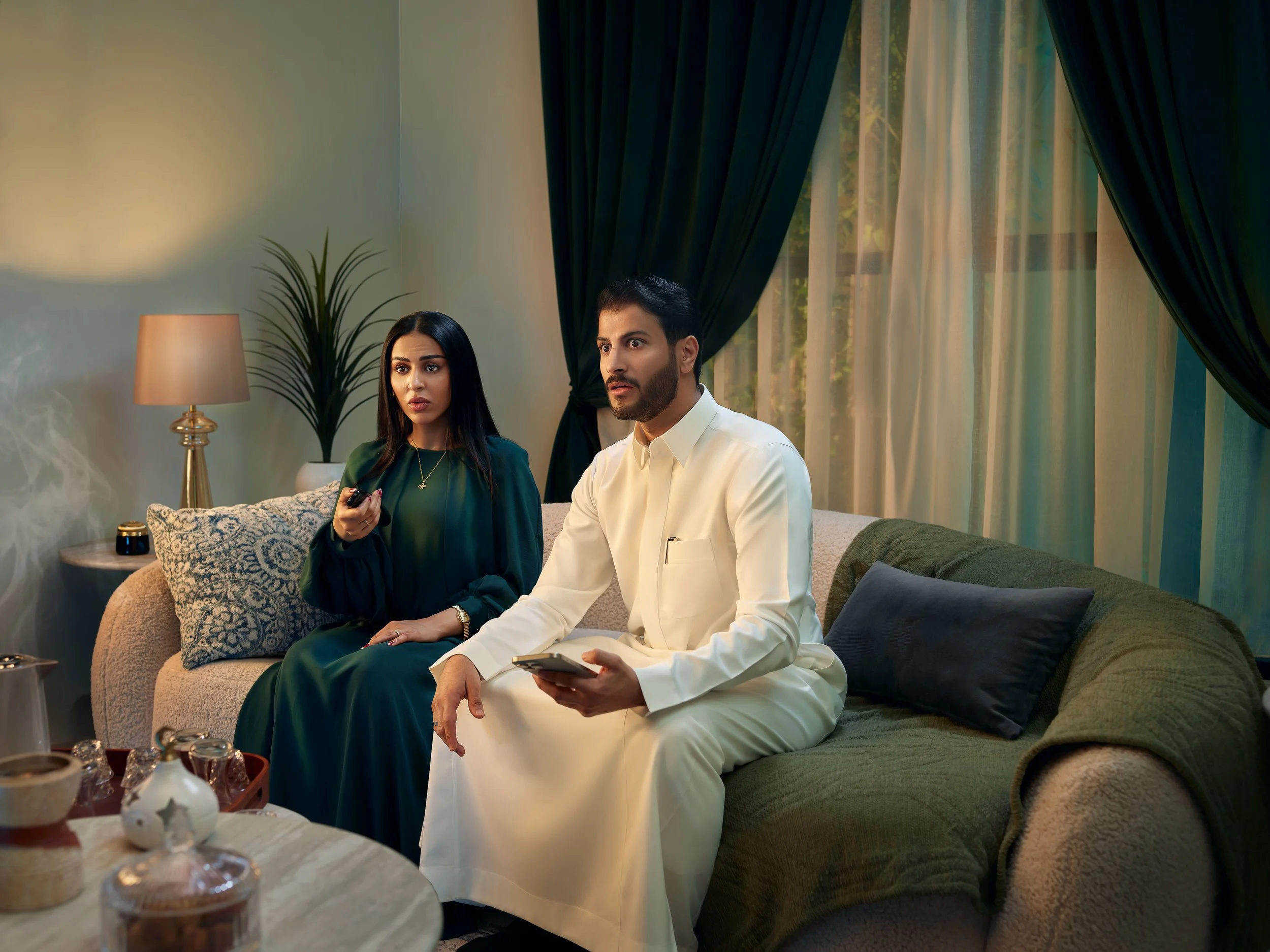 A man and woman sitting on a sofa in a living room. The man is wearing traditional white clothing and holding a remote control. The woman is wearing a dark green dress, holding a remote, and looking surprised. The room has a green sofa with pillows, 