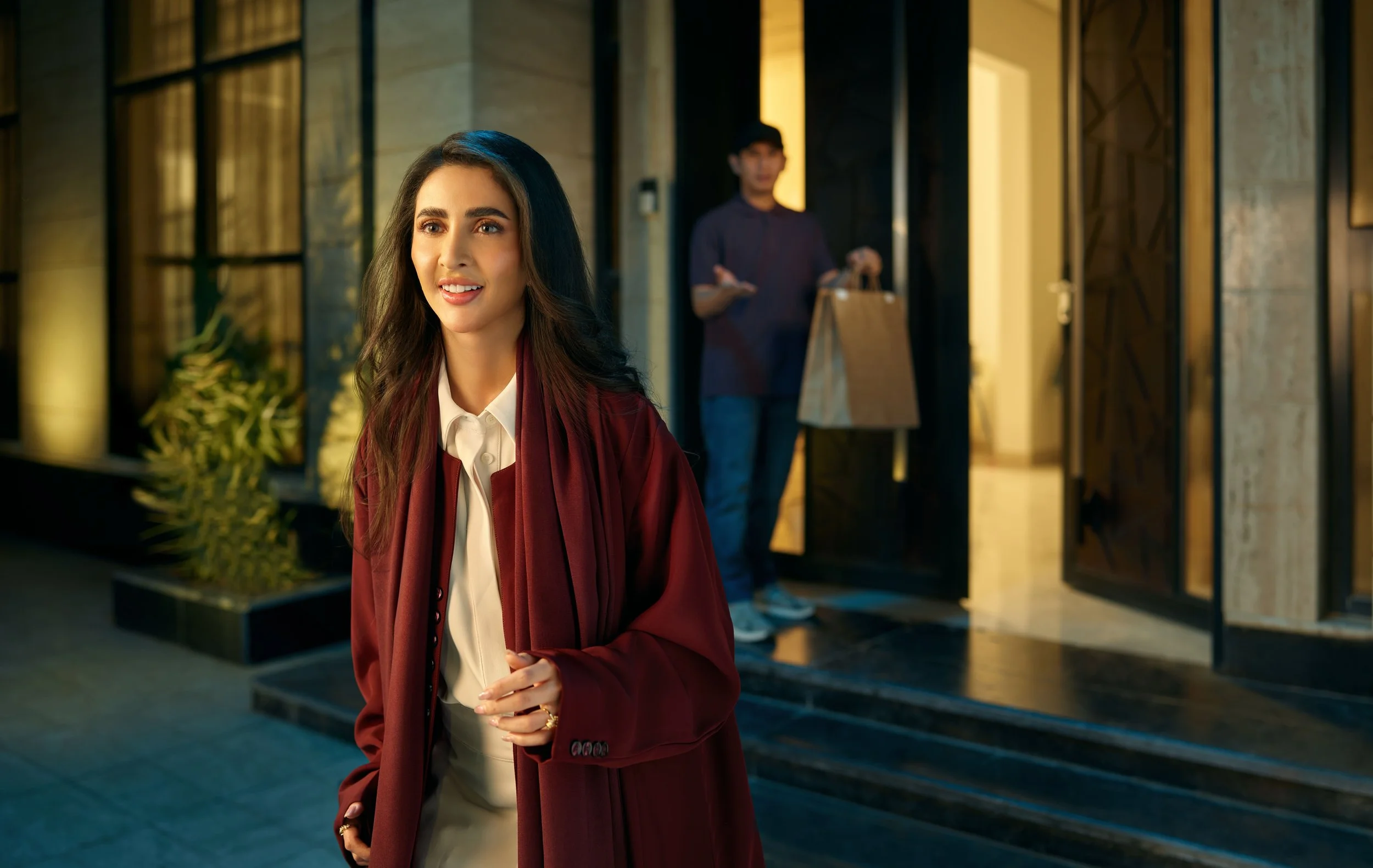 A woman with long dark hair, wearing a white shirt and a burgundy blazer, is smiling and standing outside a building at night. In the background, a man in a dark shirt and jeans is coming out of the building holding a paper bag.