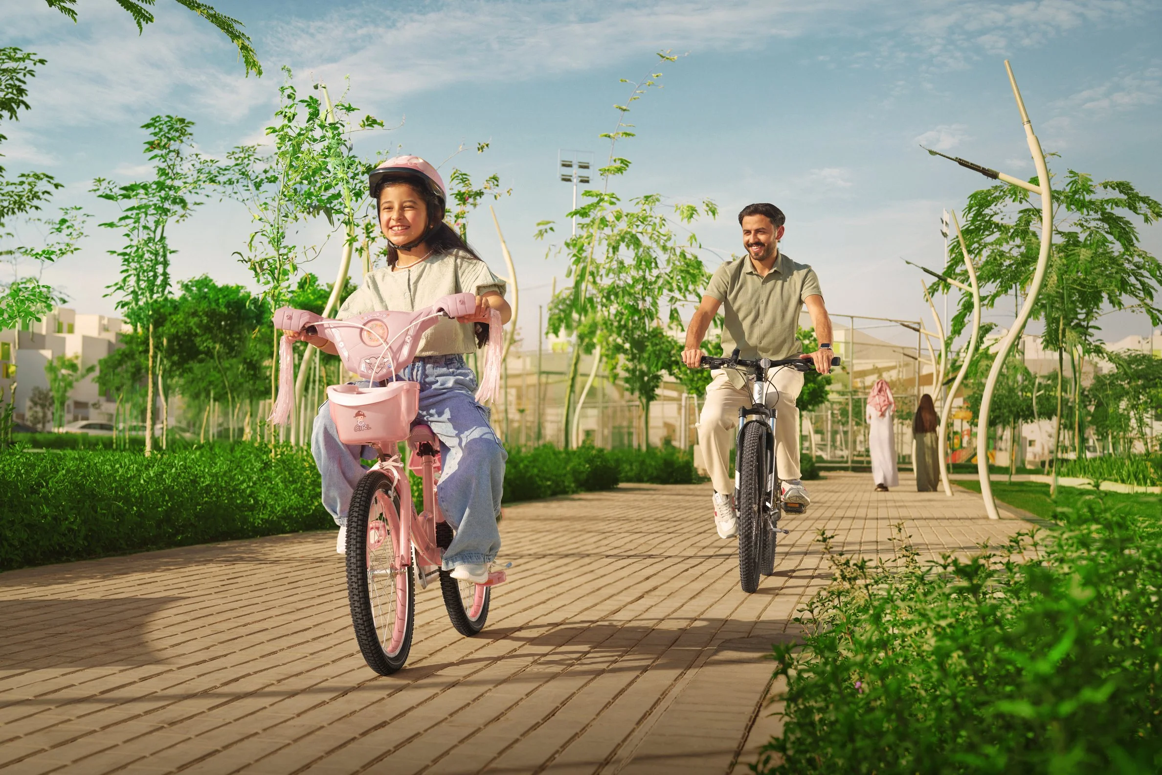 A young girl riding a pink bicycle with a helmet, and an adult man riding a black bicycle, both smiling and cycling on a paved path in a green park under a partly cloudy sky.