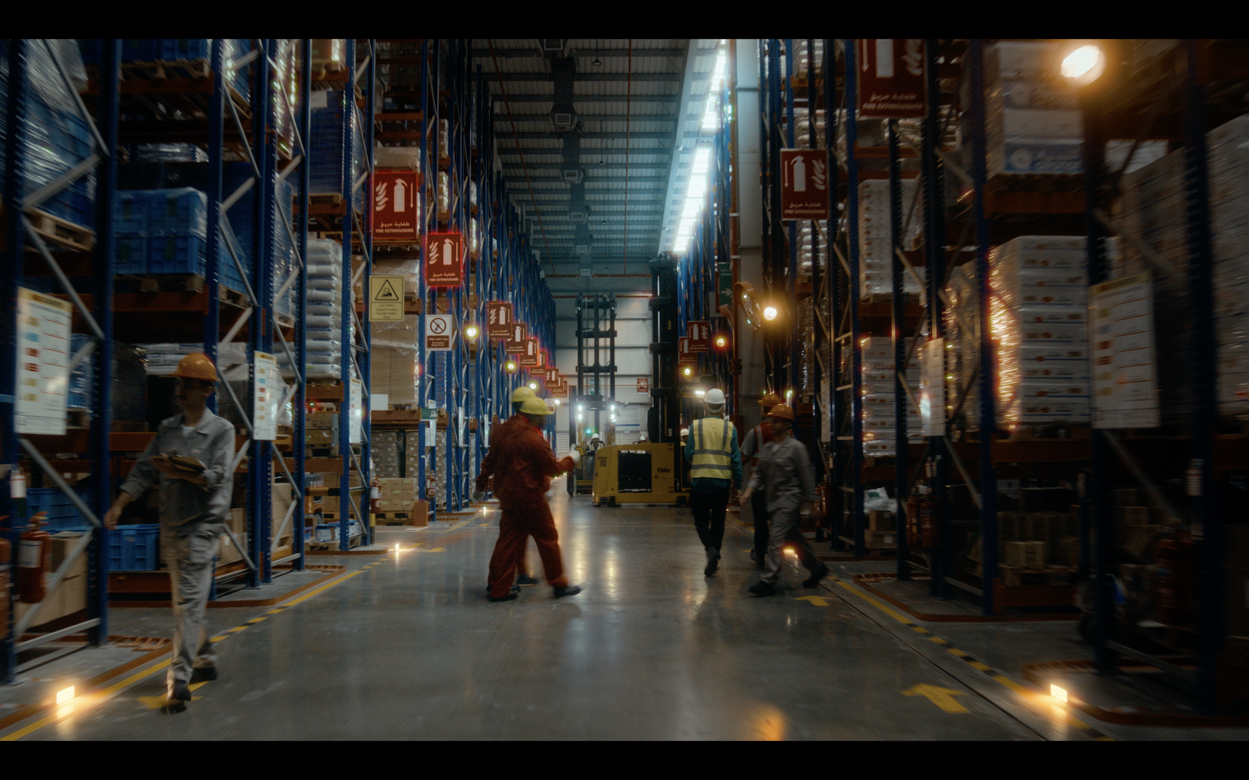 Workers in safety gear walking and inspecting in a large warehouse with tall shelves stacked with goods.