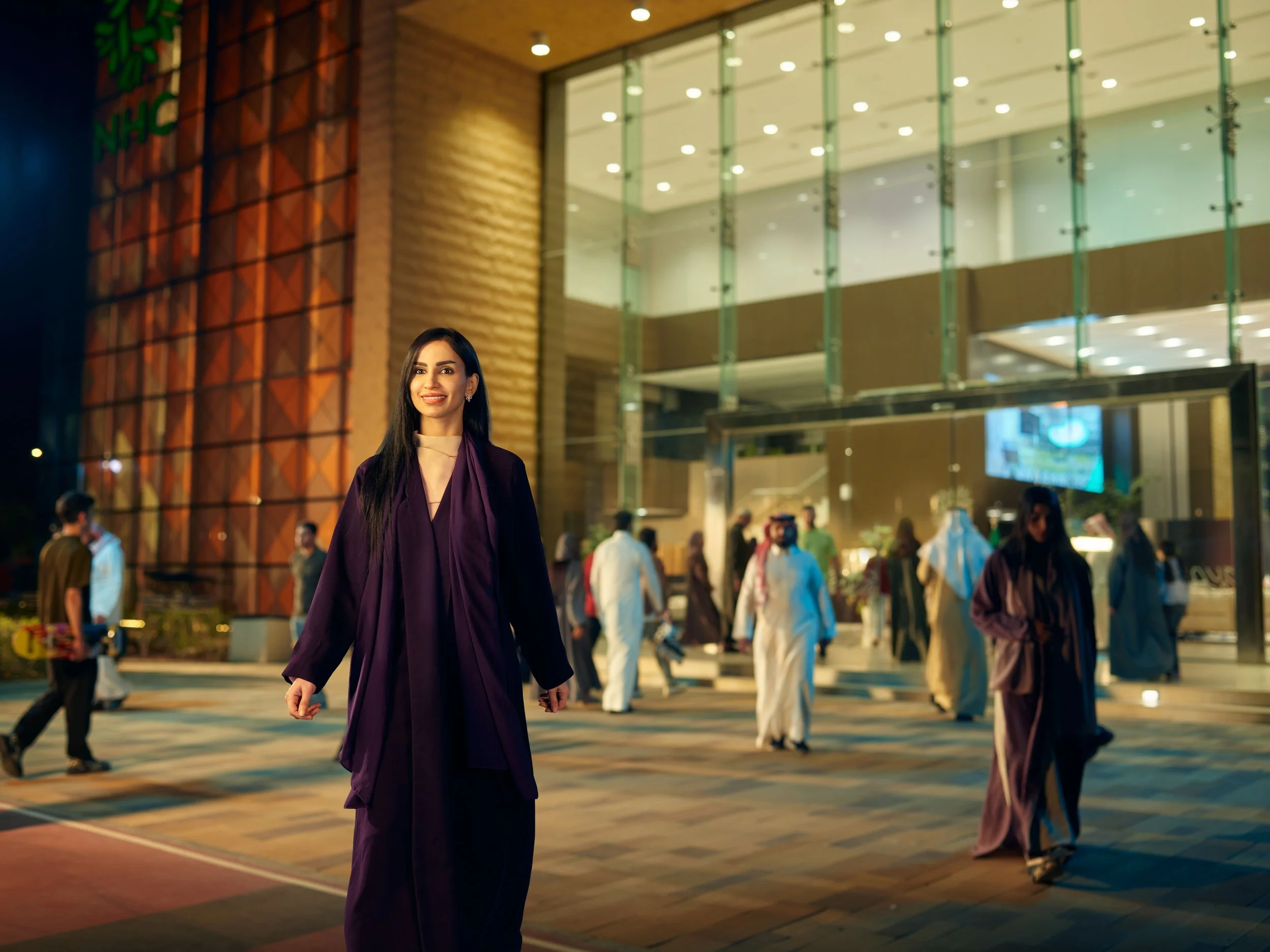 A woman in a purple dress standing outside a modern building at night, with people walking in the background.