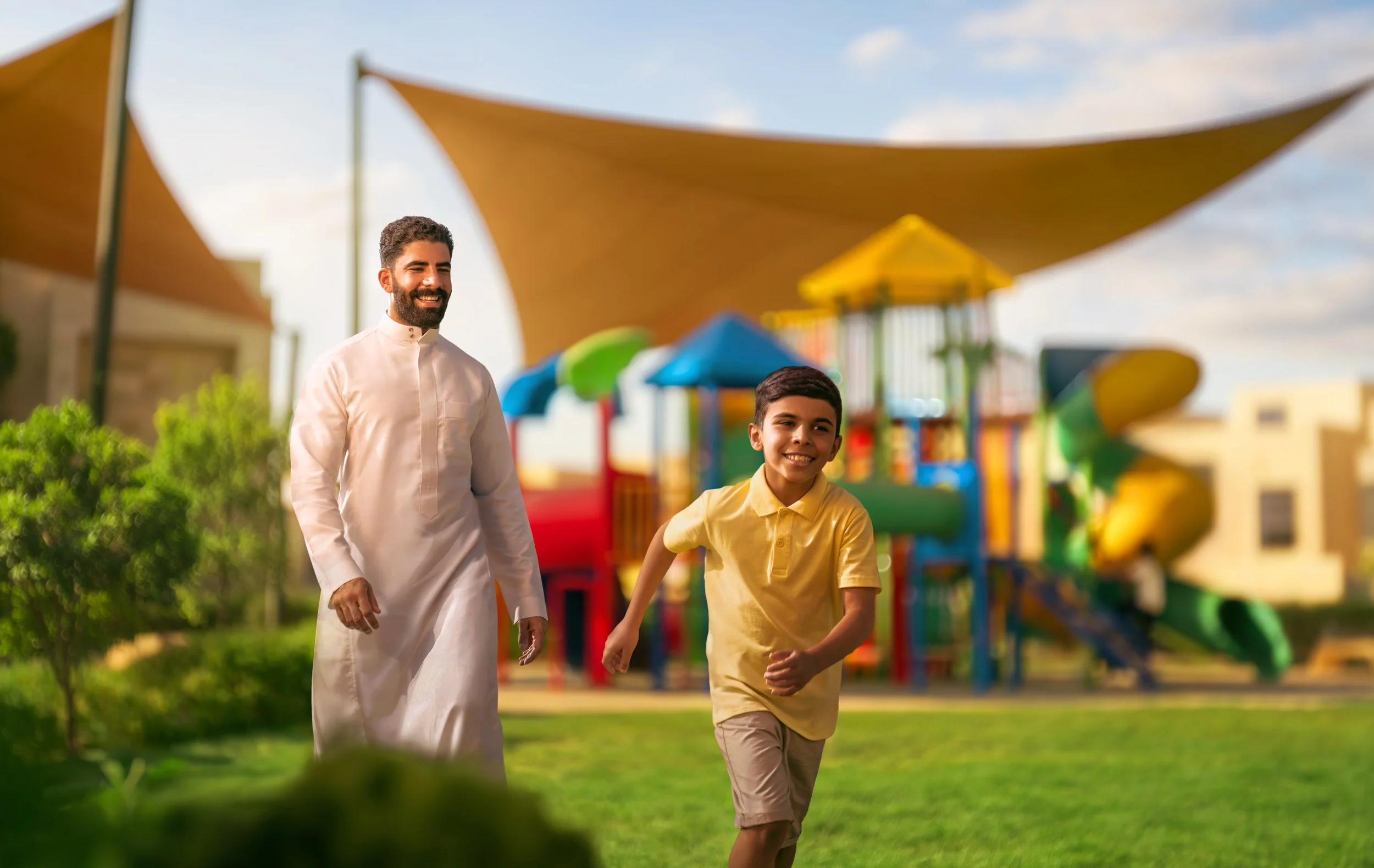 A man and a boy running and smiling in a playground with colorful slides and play structures under shade sails.