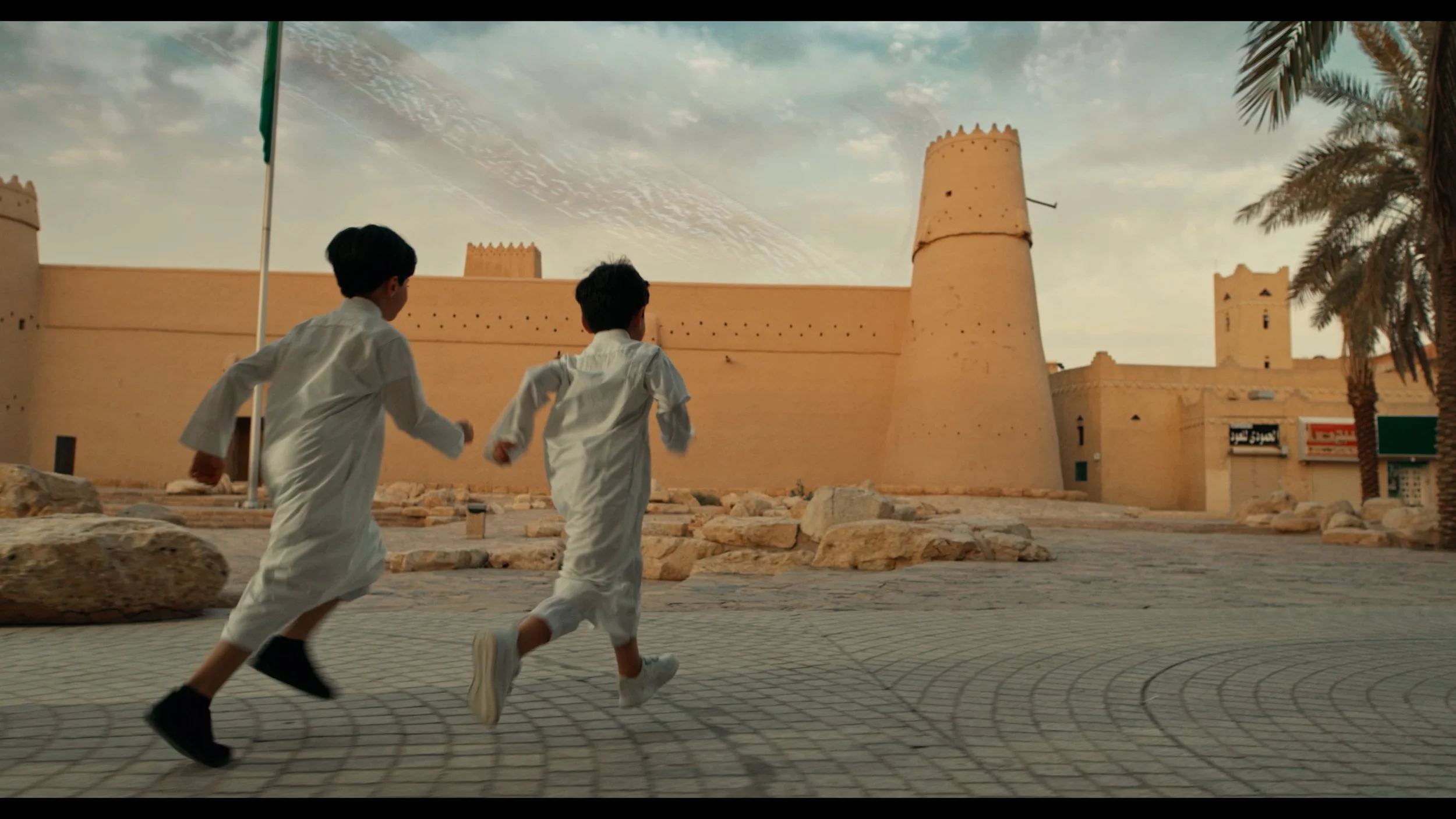 Two boys in white traditional clothing running on a paved street in front of an ancient fort or castle with beige walls, towers, and palm trees to the right, under a partly cloudy sky.