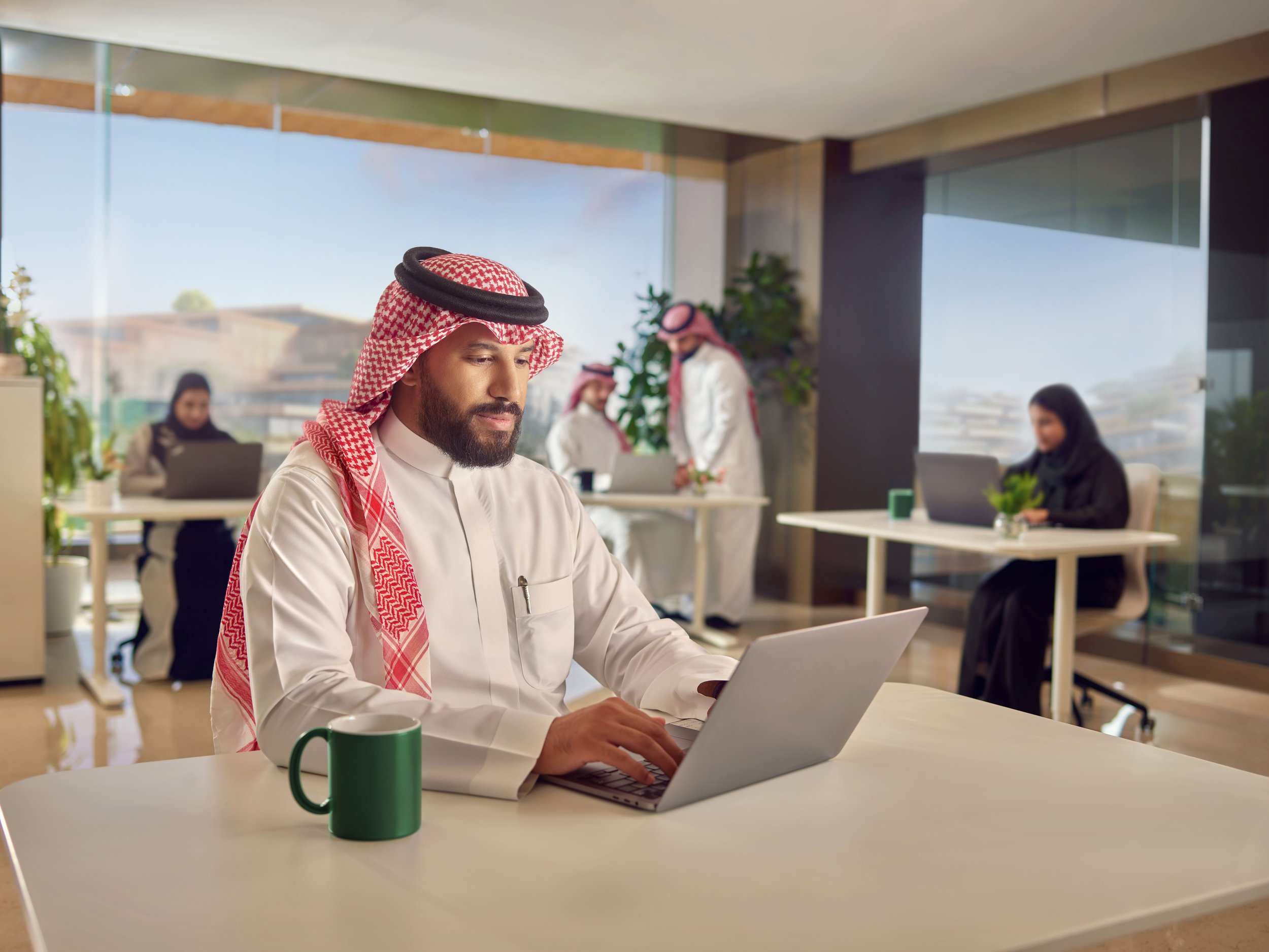 Man wearing traditional Middle Eastern attire working on a laptop at a desk in an office with other employees working in the background.