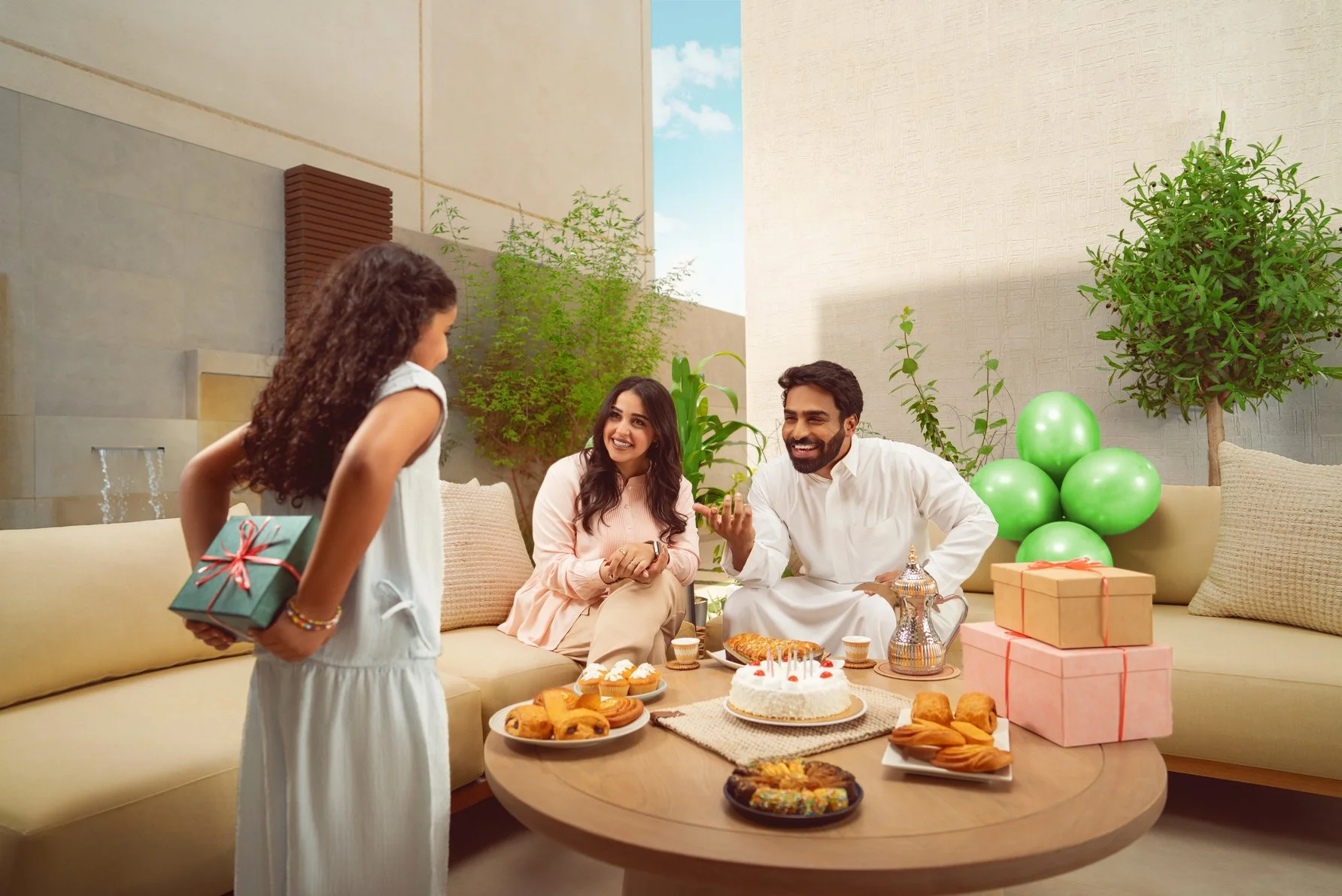 Family celebrating birthday with girl receiving a gift, cake, cookies, and balloons in a bright outdoor setting.