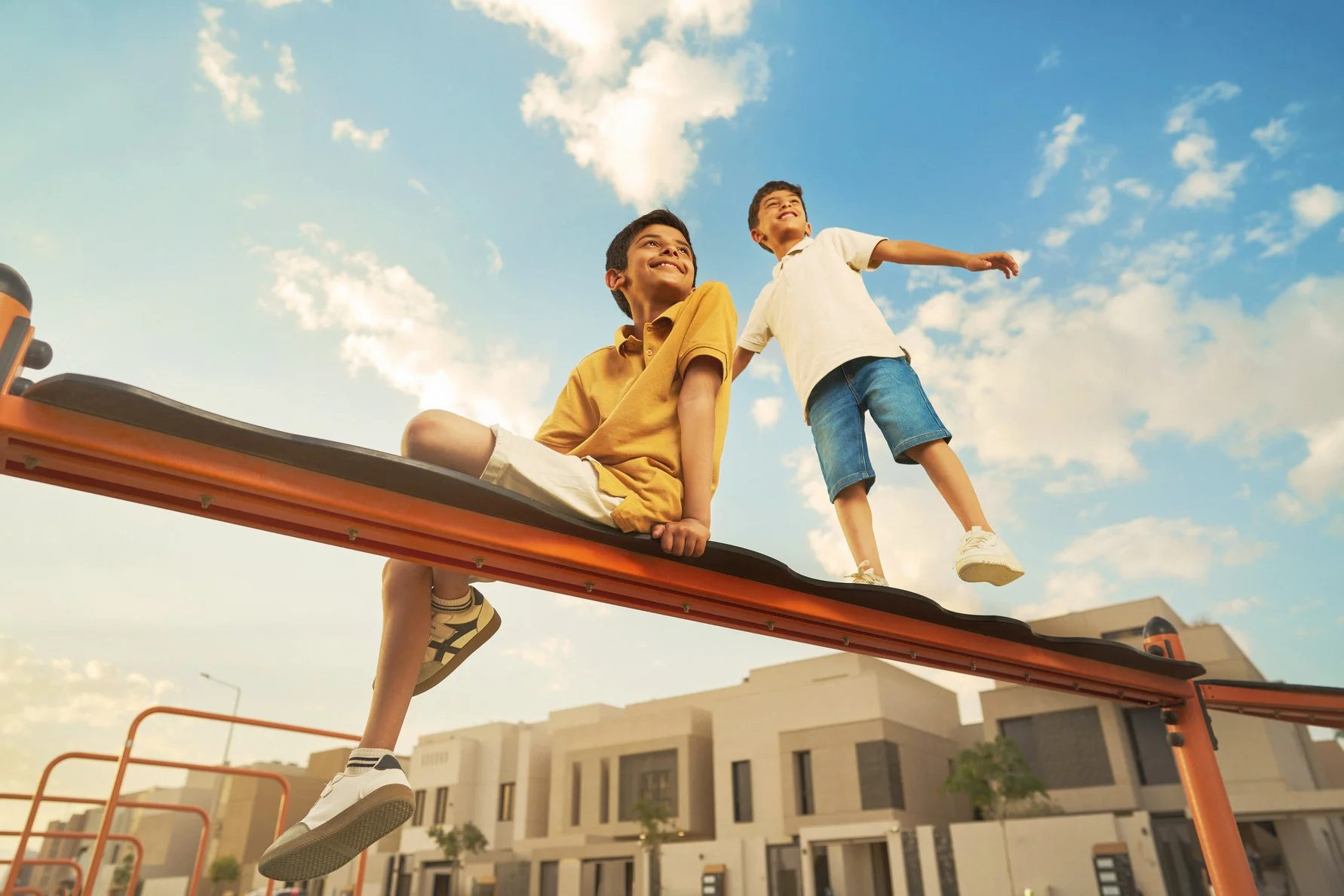 Two boys playing on a playground structure outdoors against a blue sky with scattered clouds, one sitting and smiling, the other standing and balancing with arms outstretched.
