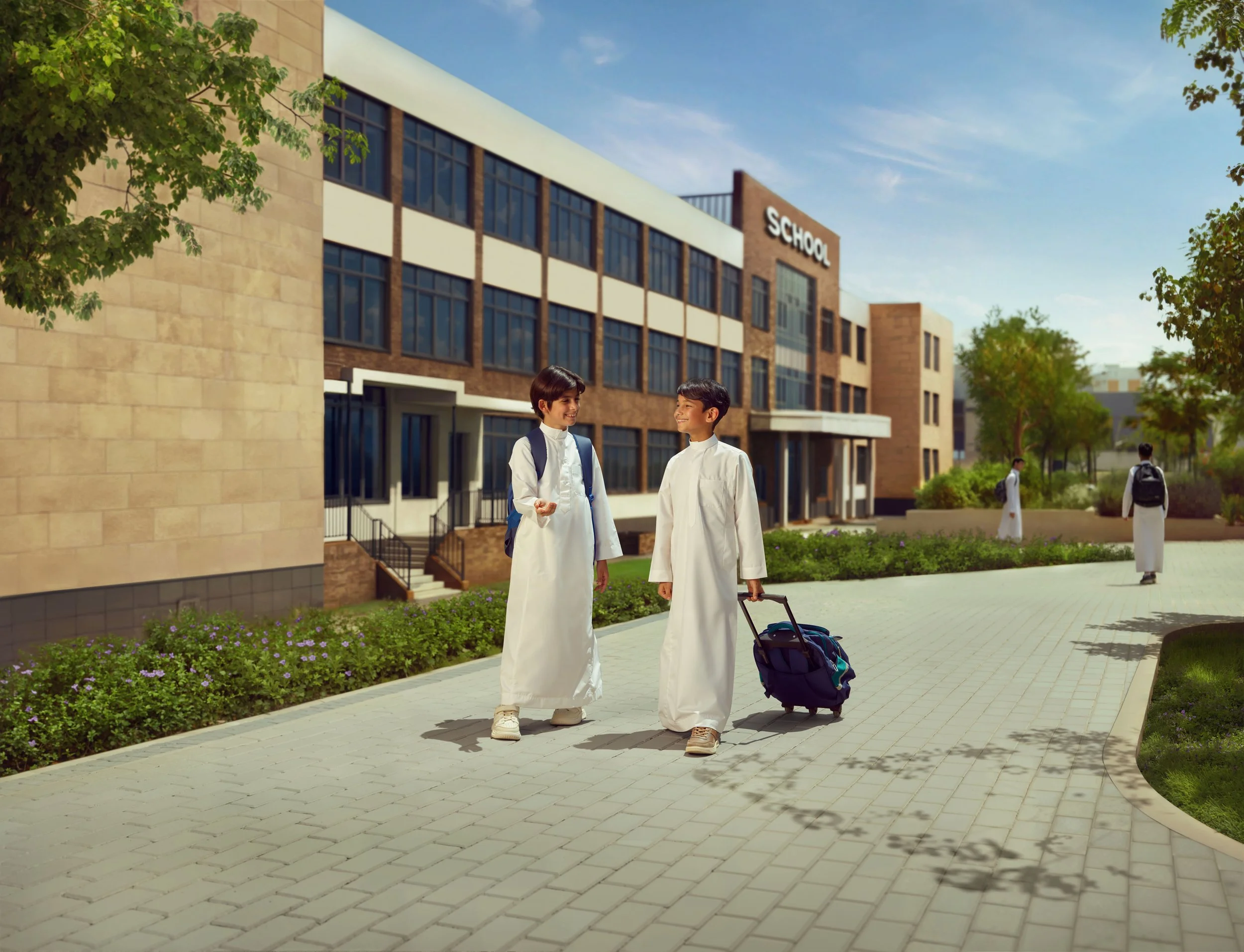 Two young boys wearing traditional white Islamic attire walking and talking outside of a modern school building on a sunny day. One boy is pulling a rolling backpack.